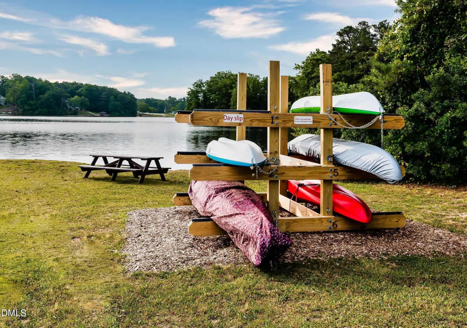 Four kayaks are stacked on a wooden rack near a lake, with a picnic table on the grass nearby. Trees and water are visible in the background under a partly cloudy sky.