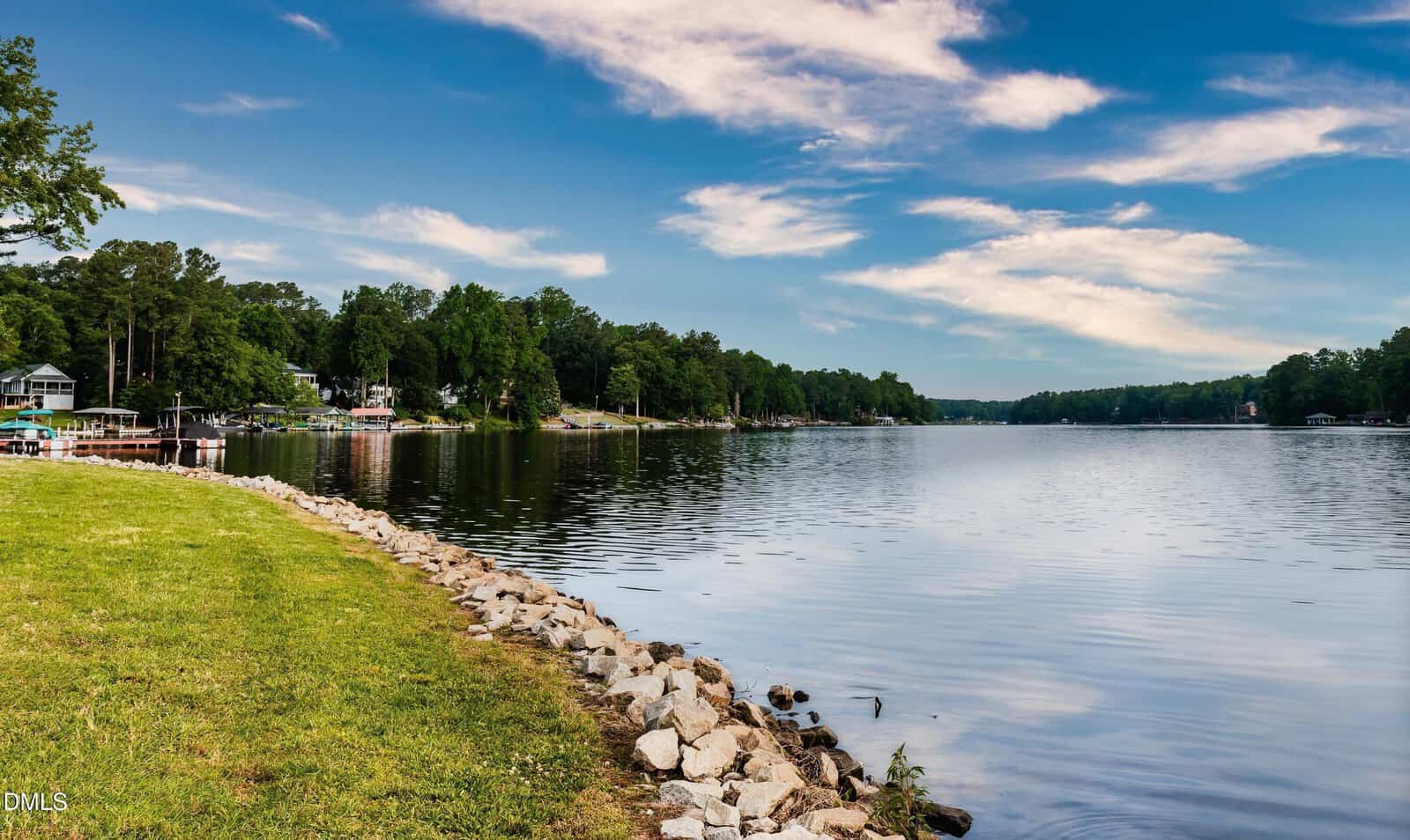 A calm lake bordered by a rocky shoreline and grassy area, with houses and trees lining the opposite shore under a partly cloudy blue sky.