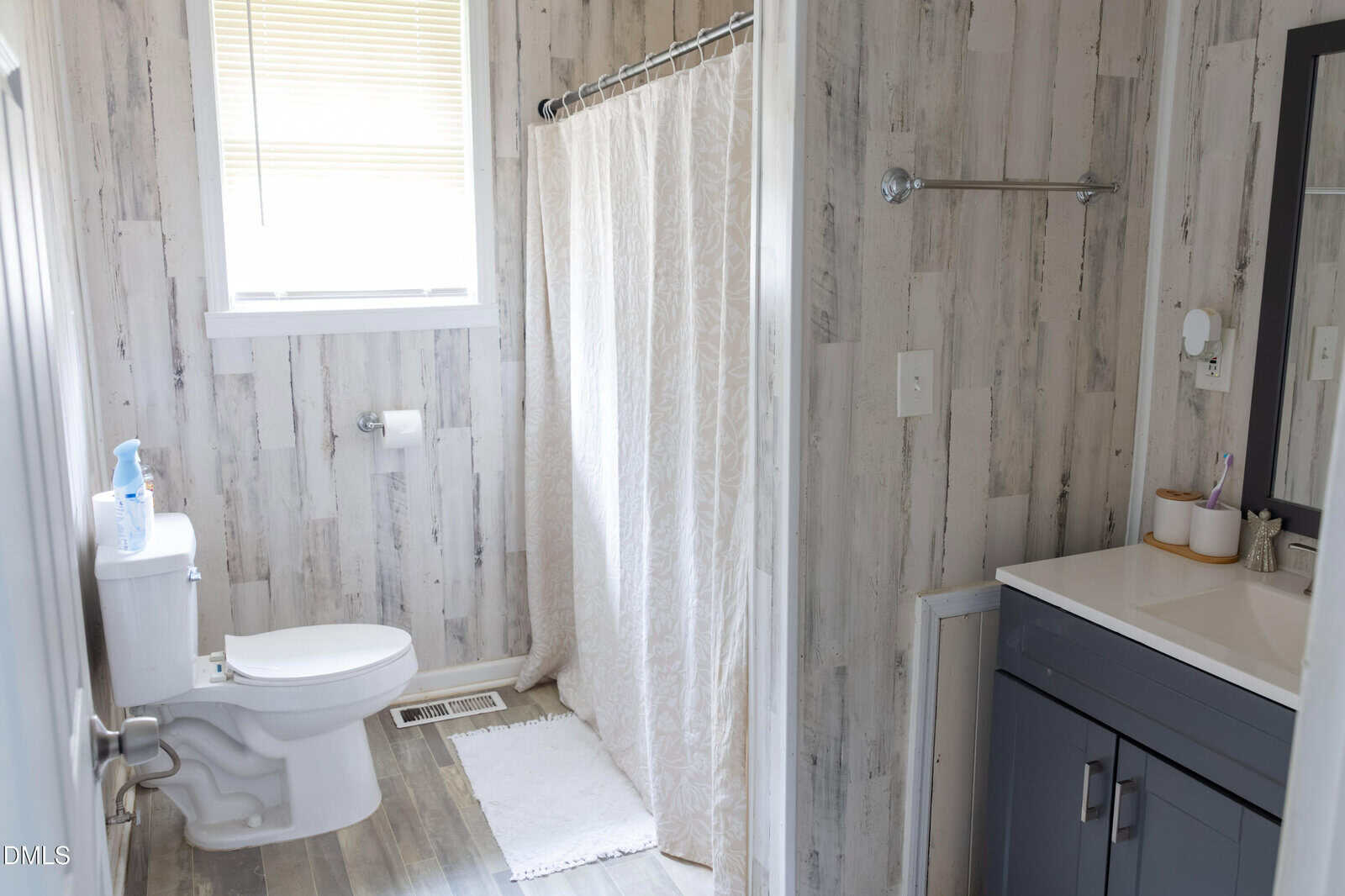 A bathroom with rustic wood walls, a white toilet, a bath mat, a shower with a curtain, a vanity with a mirror, and a window with blinds letting in natural light.