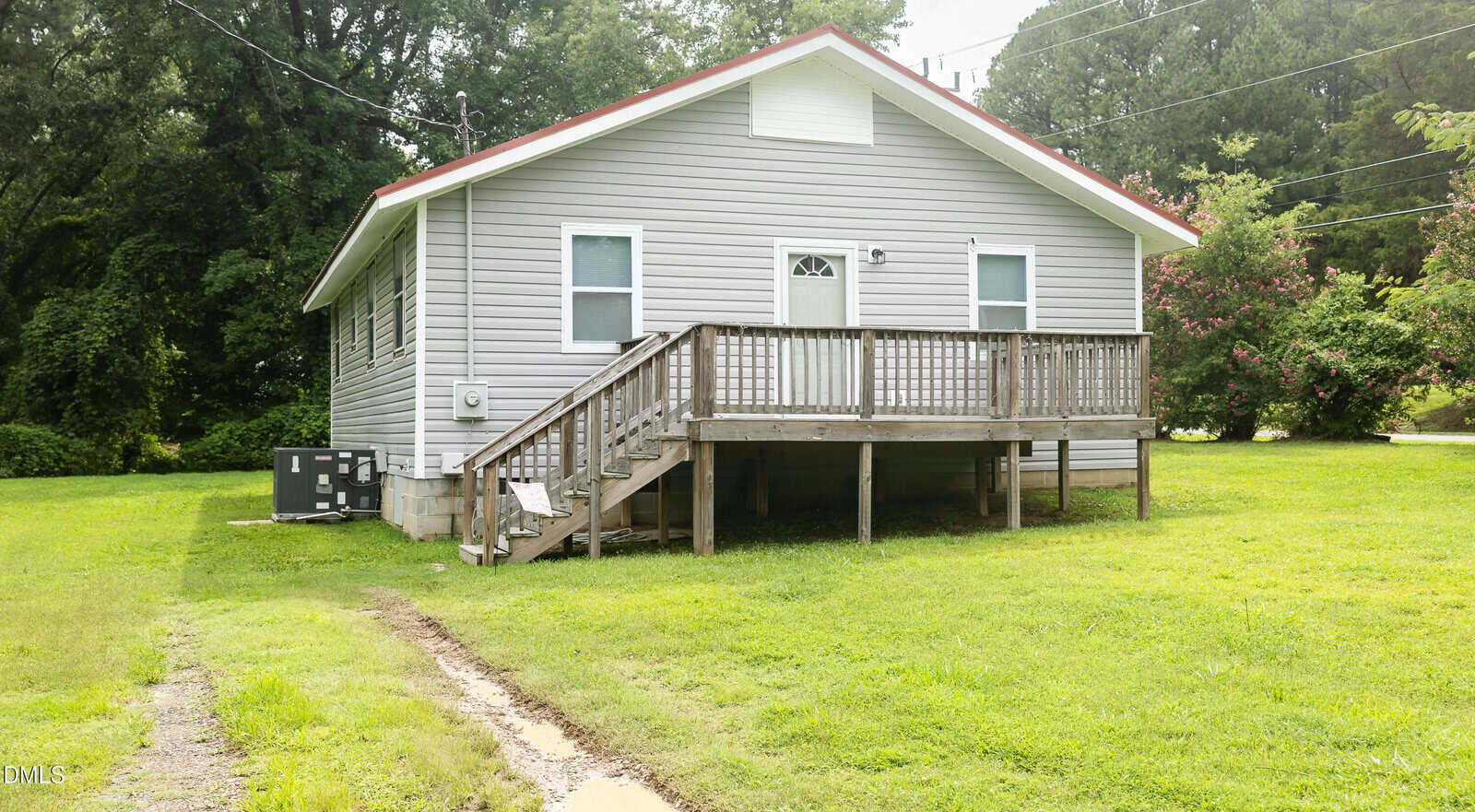 A small, light-gray house at 319 22nd Street in Butner features a front porch with steps and is elevated on concrete blocks. The yard is grassy, with a narrow dirt path leading to the porch, and trees and bushes provide a serene background.