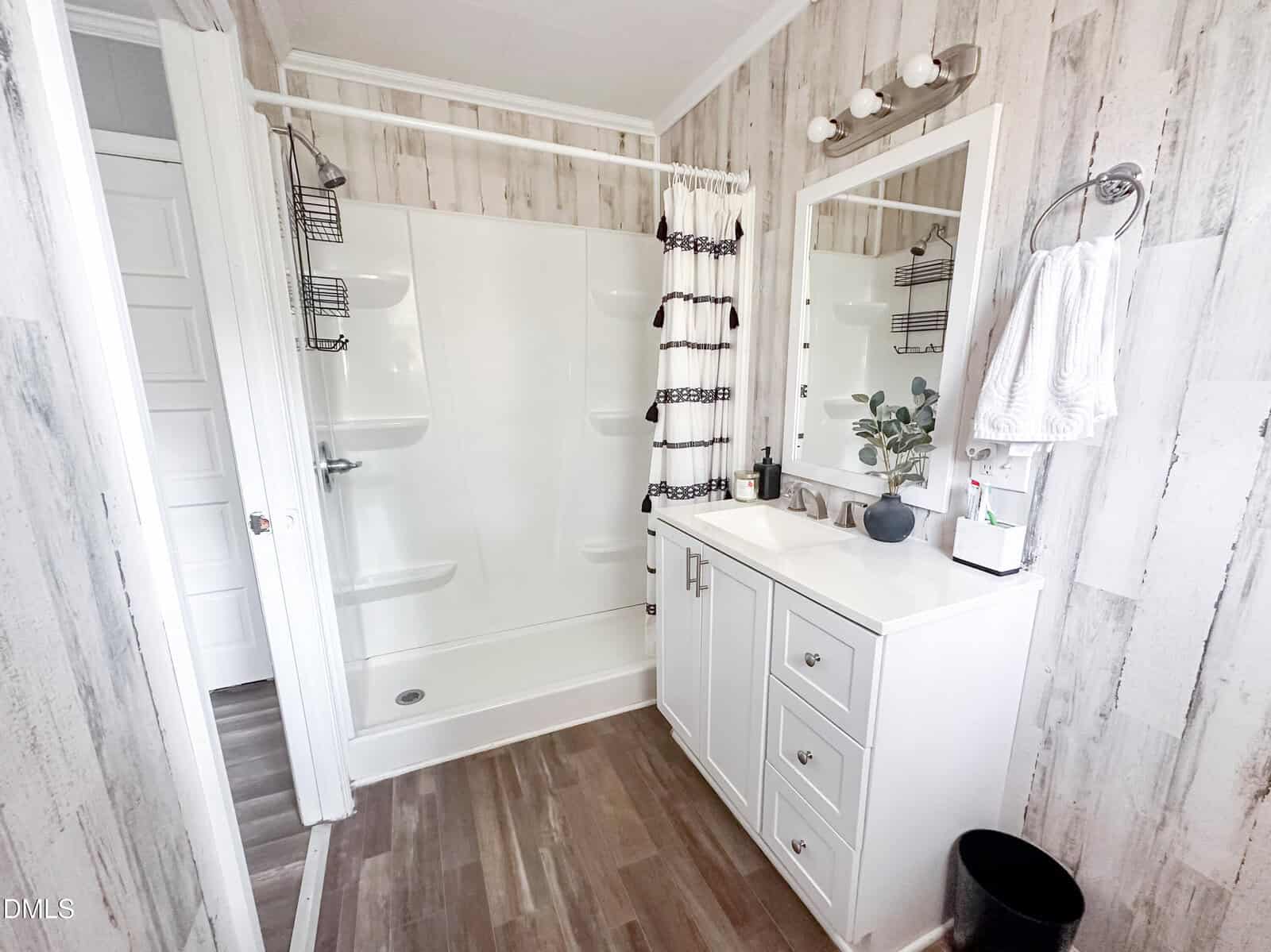 Bright bathroom with a white shower, patterned curtain, vanity with sink and mirror, towel on the wall, soap dispensers, and a plant. Light wood-look flooring and wood-pattern walls. Door visible to the left.