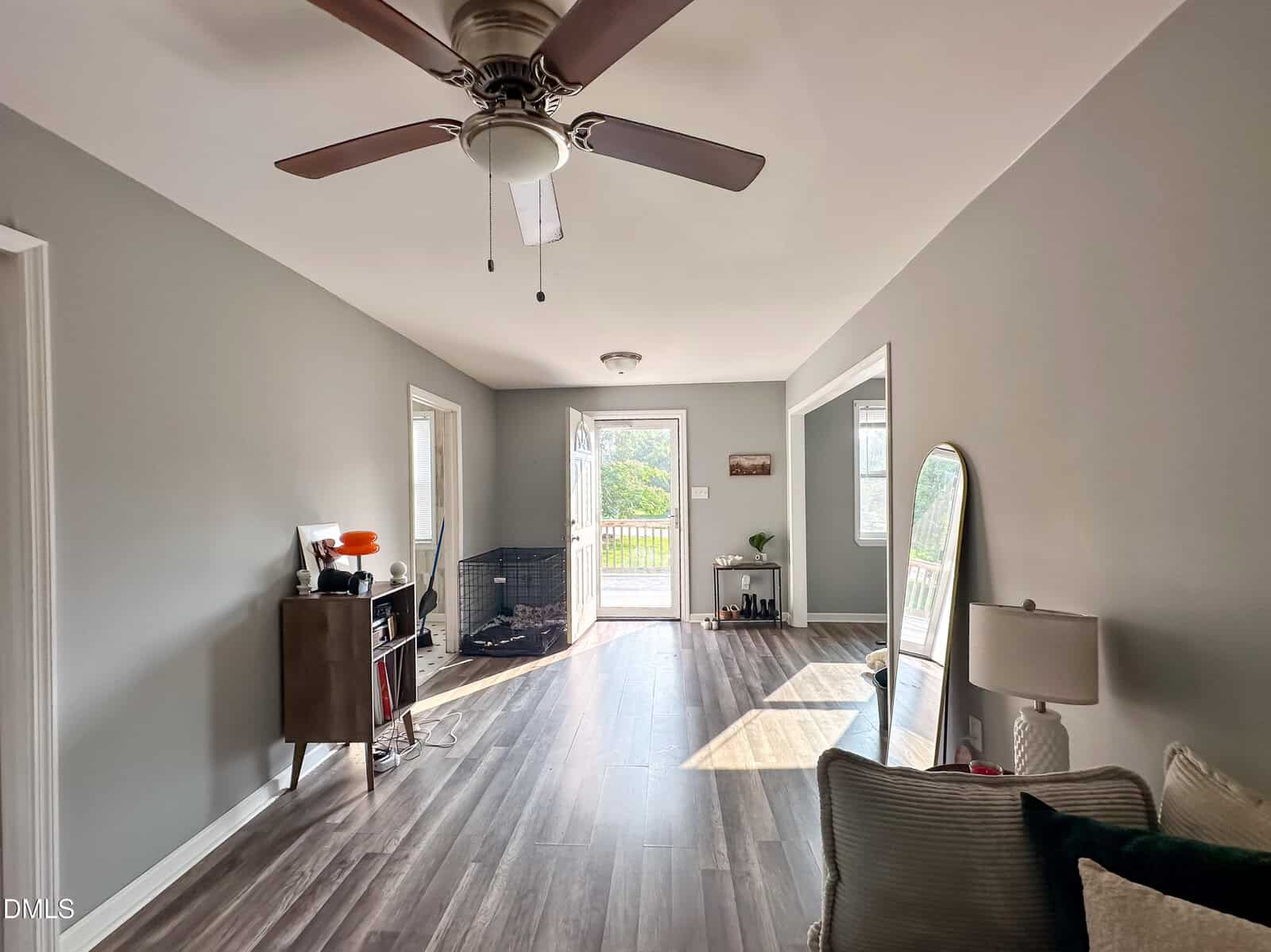 A bright, modern living room with gray walls, wood flooring, a ceiling fan, a mirror, a small cabinet, a lamp, and an open glass door leading to a deck with greenery outside.