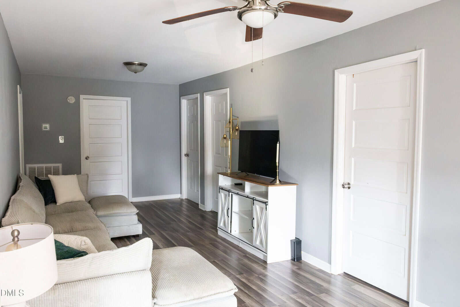 A living room with gray walls, wood flooring, a beige sectional sofa, a white TV stand with a flat-screen TV, a lamp, and three white doors. A ceiling fan with lights is mounted on the ceiling.