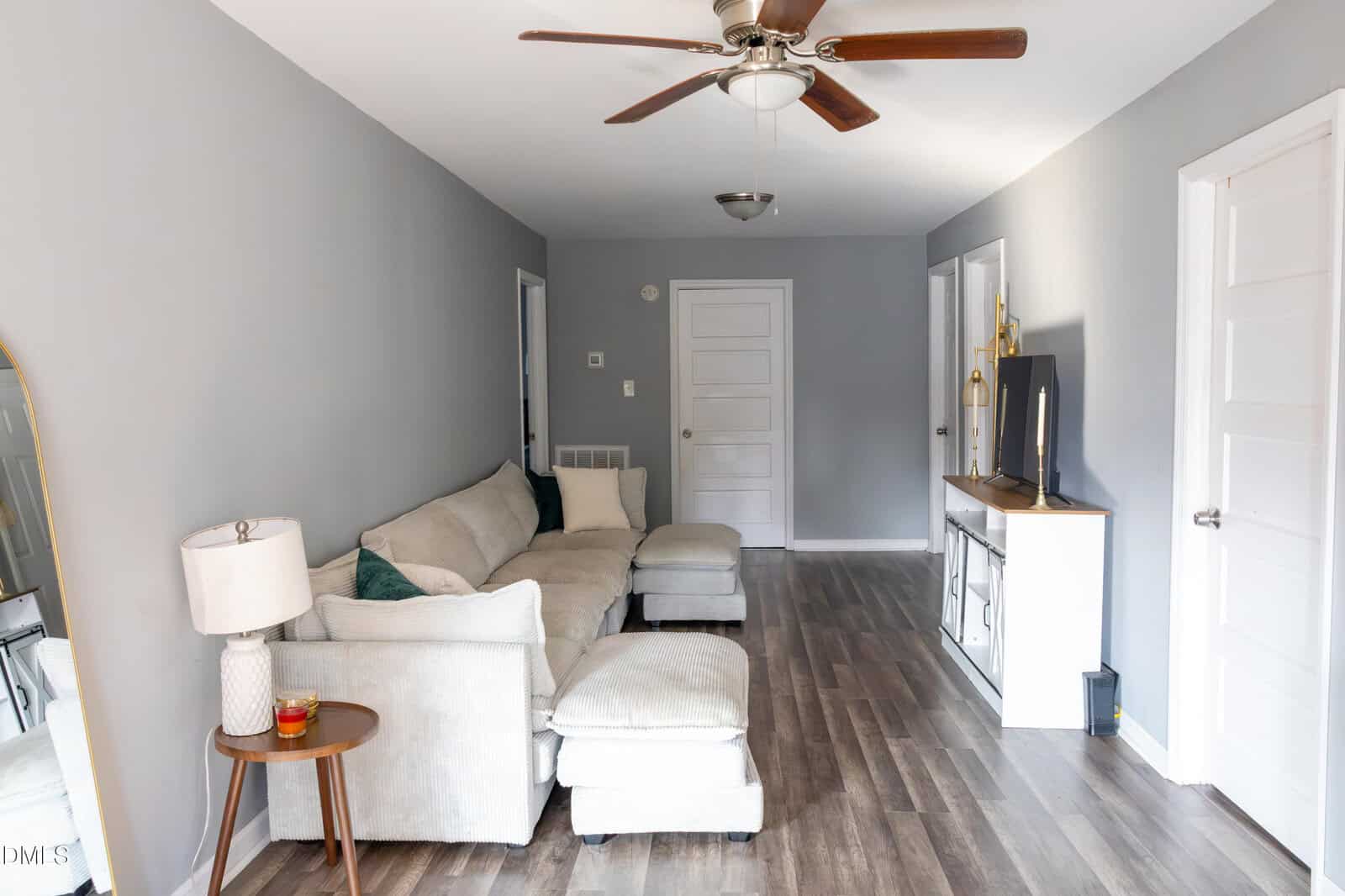 A living room with gray walls, a beige sectional sofa, a wooden side table with a lamp and candle, a TV on a console, wood-look flooring, and a ceiling fan with wooden blades.