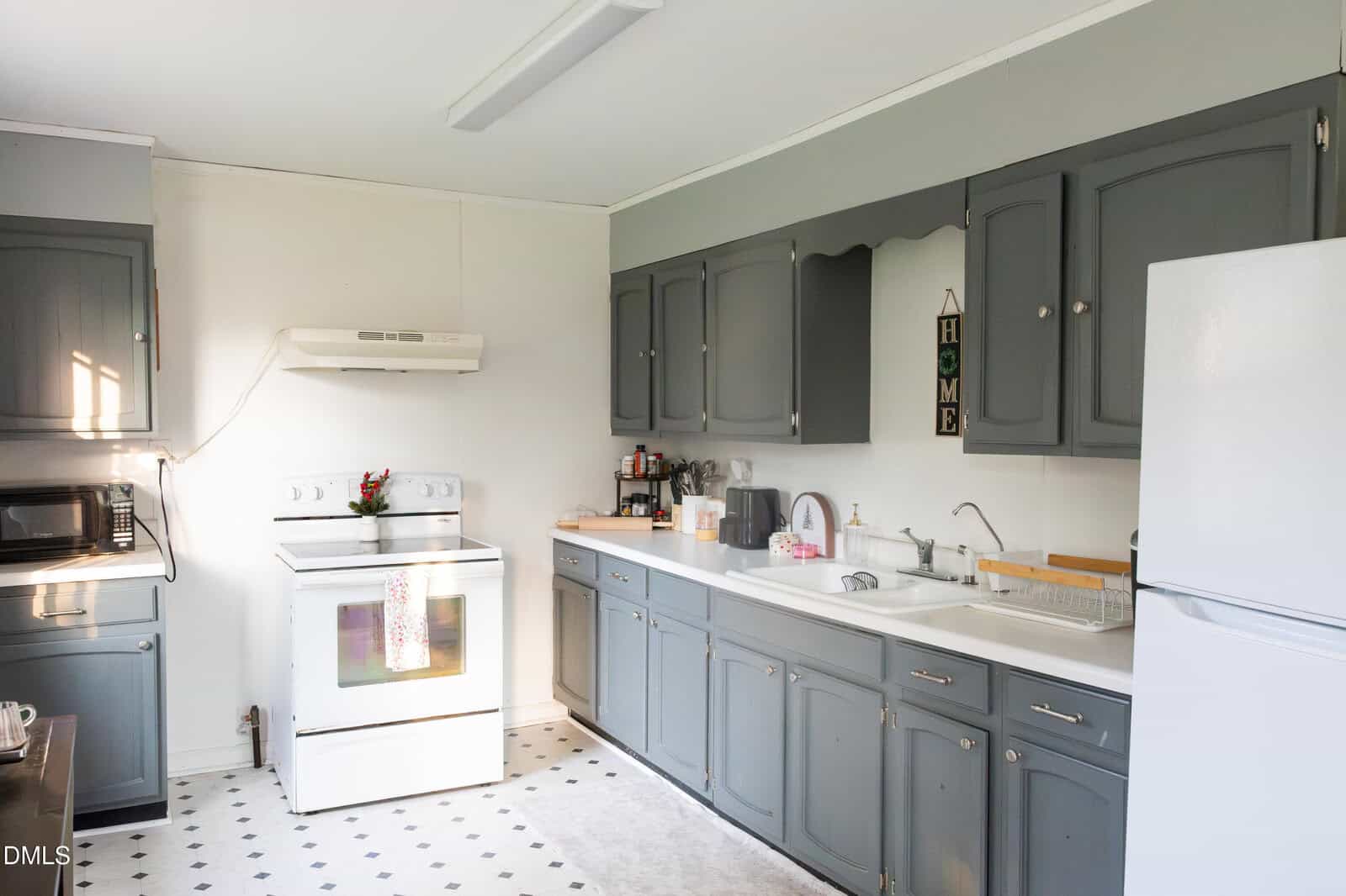 A kitchen with gray cabinets, a white stove, a white fridge, a microwave, a double sink, and various kitchen items on the counters. Light comes in from a window on the left. The floor is white with a small pattern.