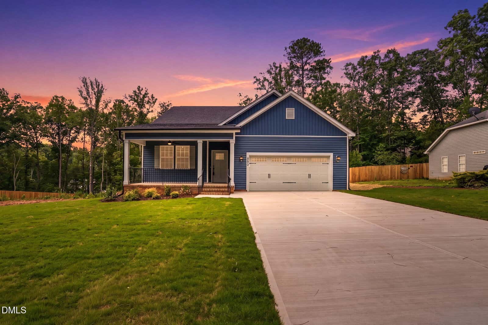 A blue single-story house with a covered front porch, white trim, and a two-car garage, set against a sunset sky. The home has a large driveway and a green lawn, with trees and a wooden fence in the background.
