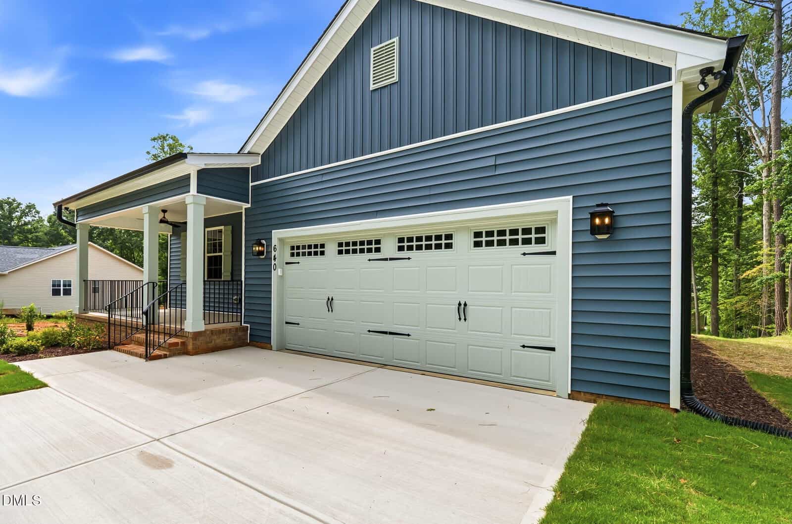 A modern house with blue siding, white trim, a large three-car garage door, front porch with black railings, brick steps, and a concrete driveway. Green lawn and trees surround the property.