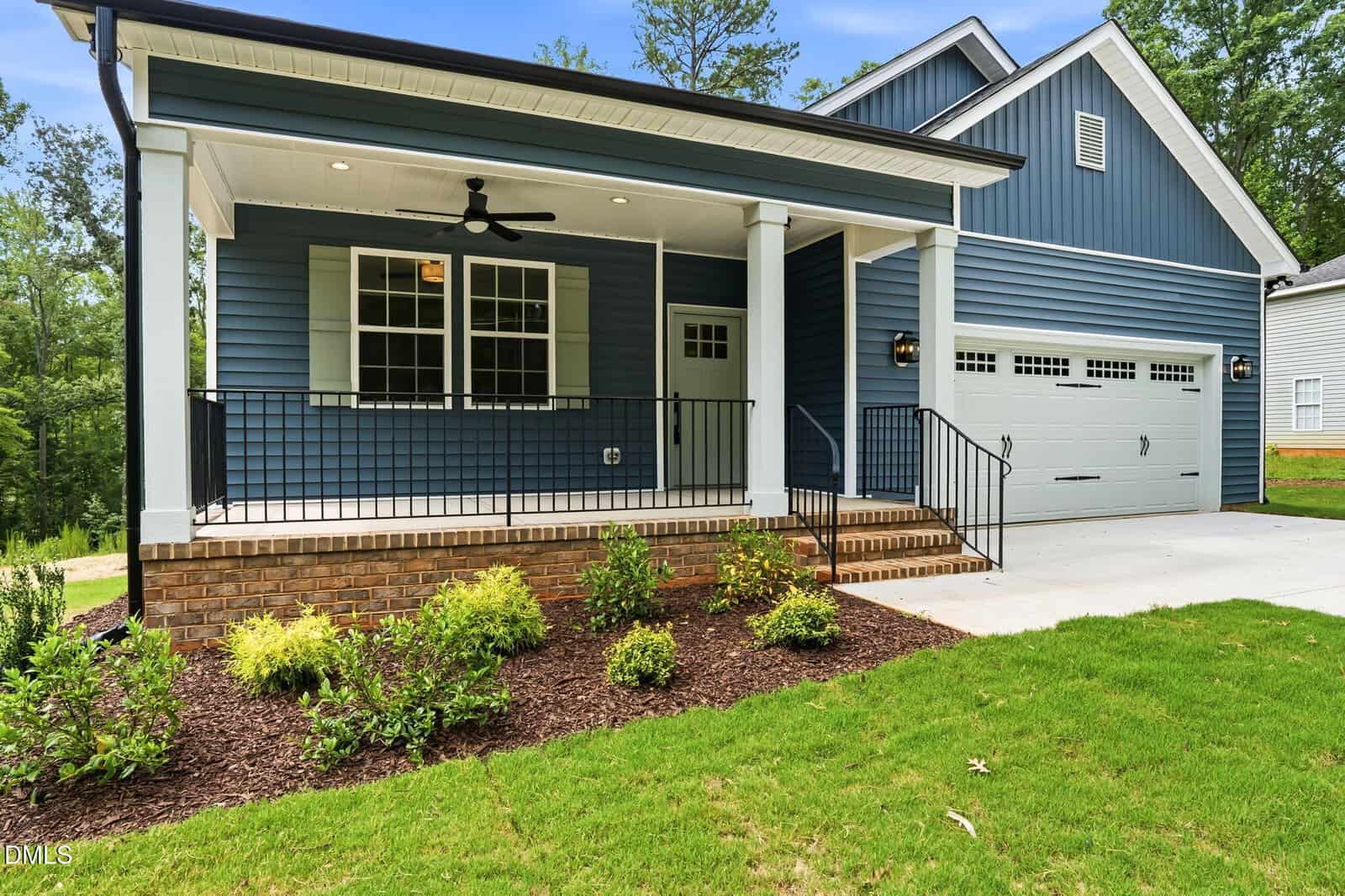A blue single-story house with a covered front porch, white trim, black railings, a ceiling fan, two-car garage, brick foundation, landscaped yard with shrubs, and a concrete driveway.
