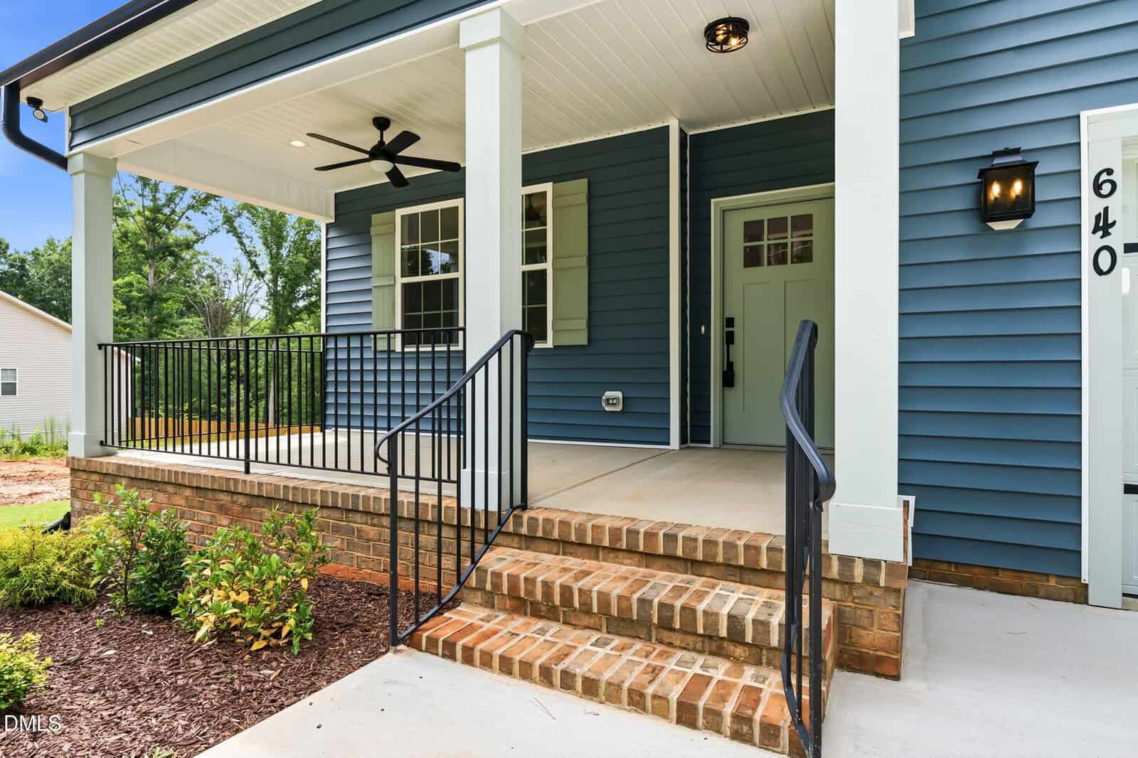 Front porch of a house with blue siding, white trim, brick steps, black metal railings, a ceiling fan, and a mint green front door. There is a porch light and house number 640 displayed on a column.