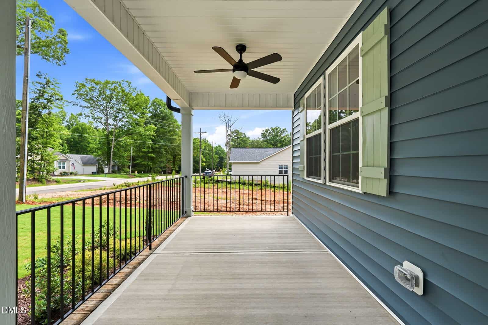 Covered front porch with black metal railing, ceiling fan, and blue siding. Large windows overlook a green yard and quiet suburban street with houses and trees in the background.