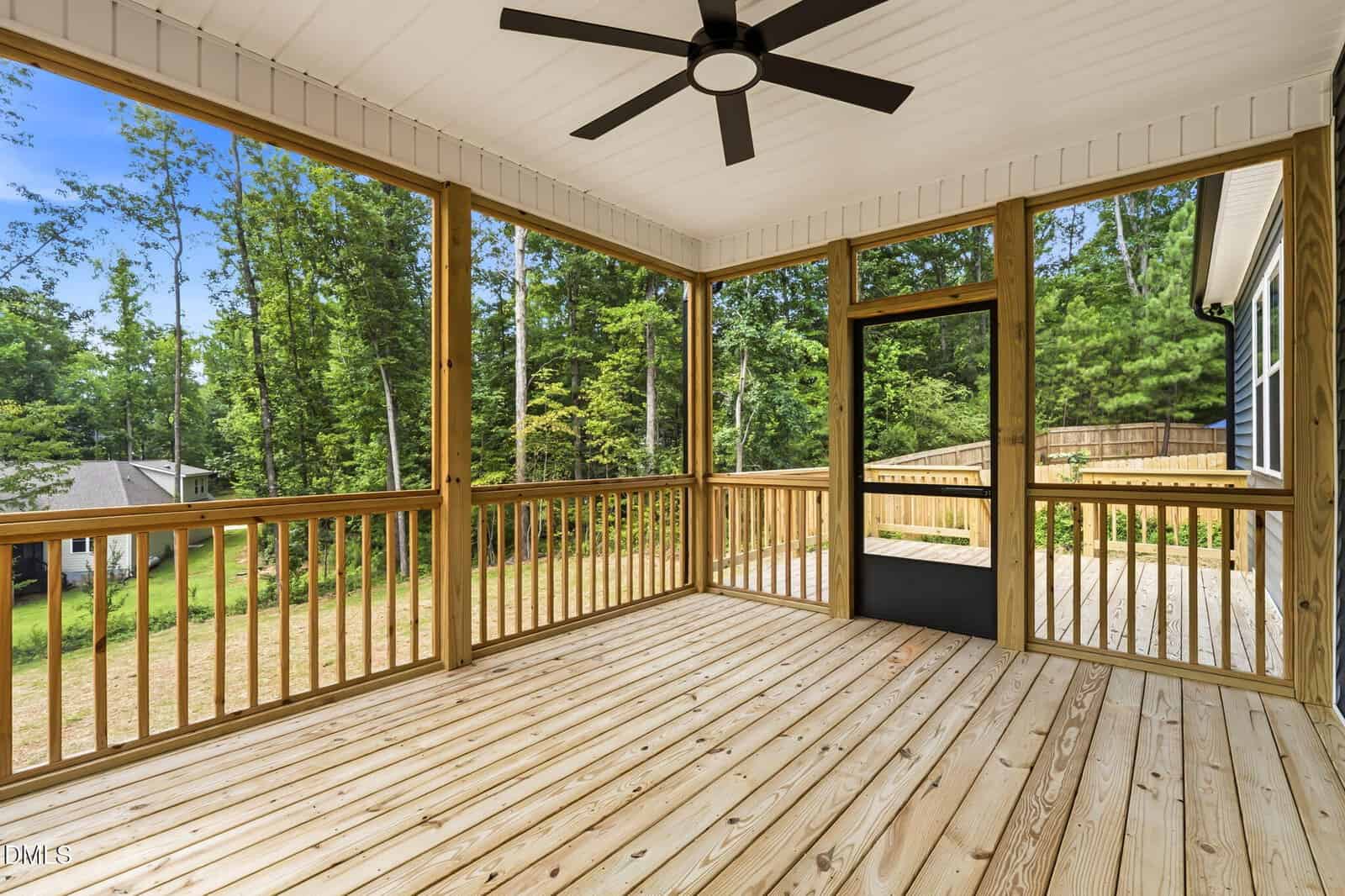A screened-in wooden porch with a ceiling fan, surrounded by trees and overlooking a grassy yard, attached to a house.