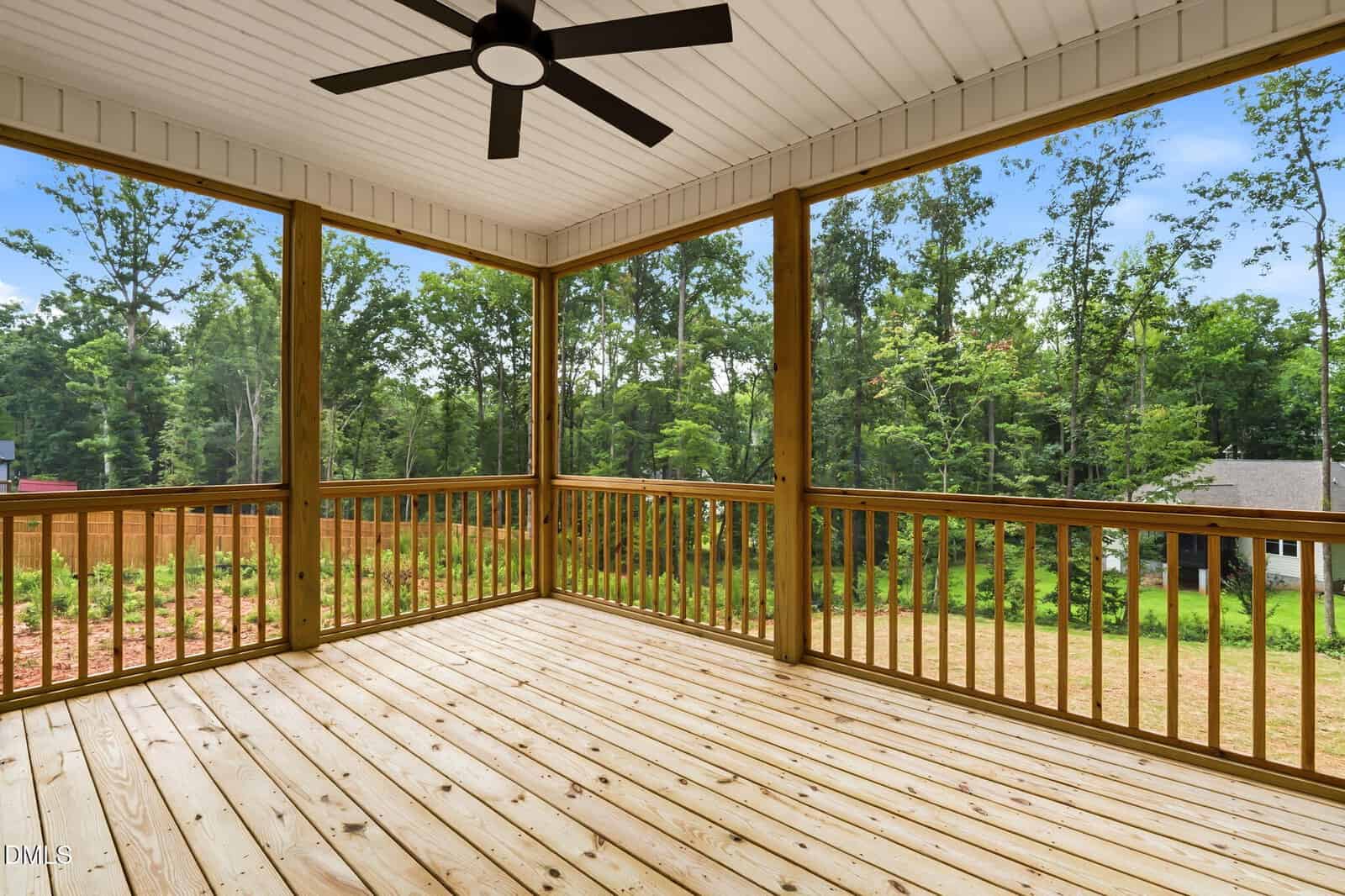 Screened-in wooden porch with a ceiling fan, surrounded by a wooden railing, overlooking a green backyard and forested area under a blue sky.