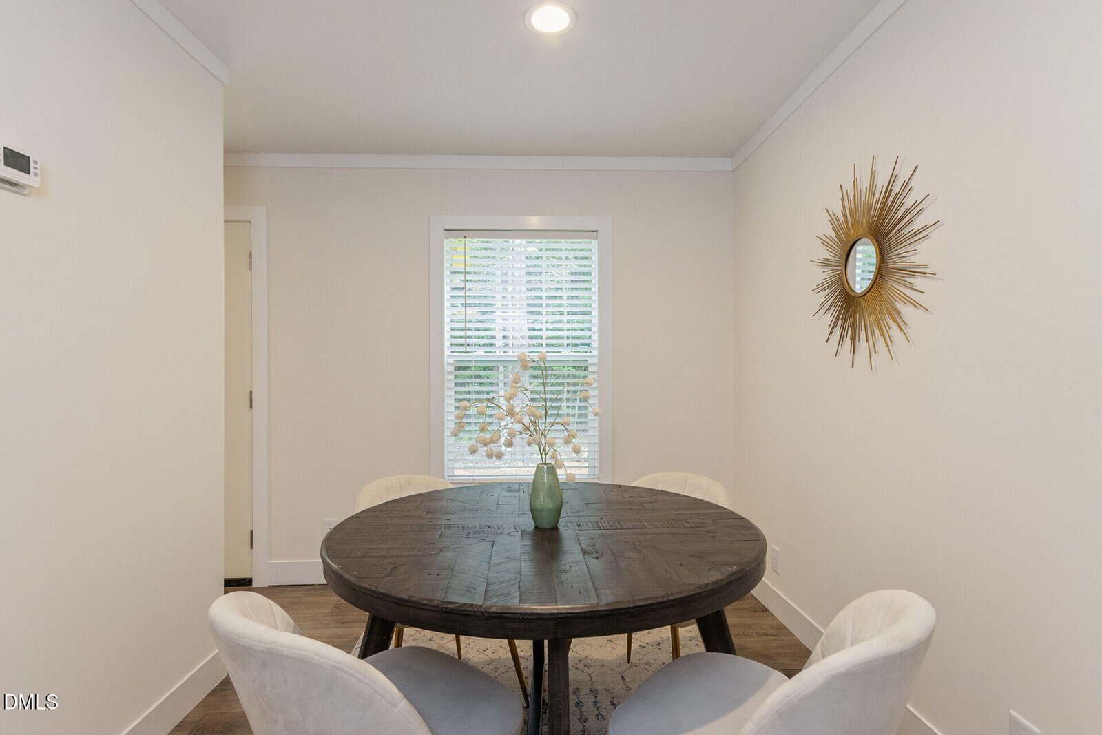 Small dining room with a round dark wood table, four white chairs, a green vase with white flowers, a window with blinds, and a sunburst mirror on the right wall.