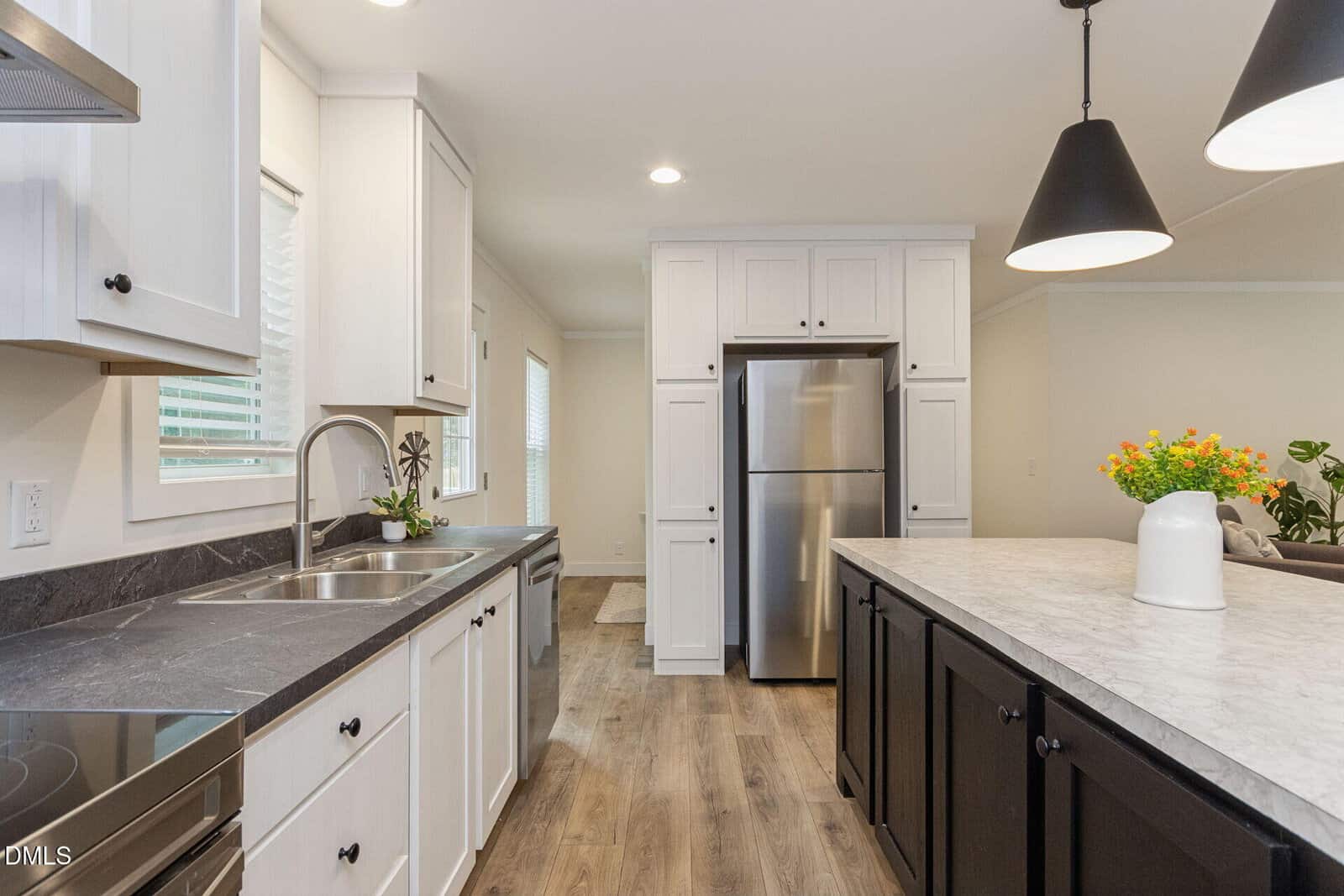 Modern kitchen with white cabinets, stainless steel refrigerator, black countertops, large island with marble surface, two black pendant lights, wood flooring, and decorative plants by the sink and on the island.
