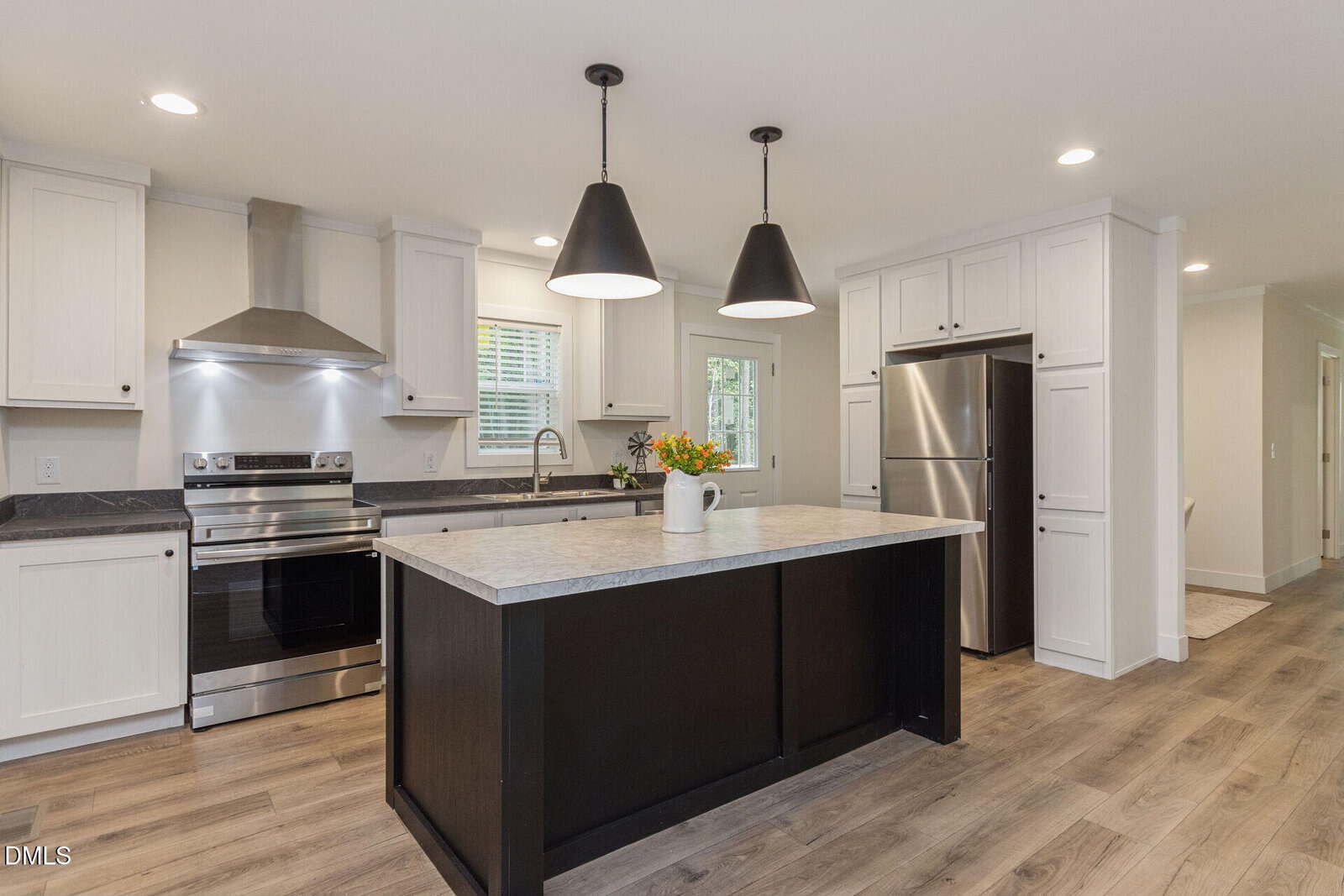 Modern kitchen with white cabinets, stainless steel appliances, a center island with a marble countertop, black pendant lights, wood flooring, and a vase with flowers on the island. A window is above the sink.
