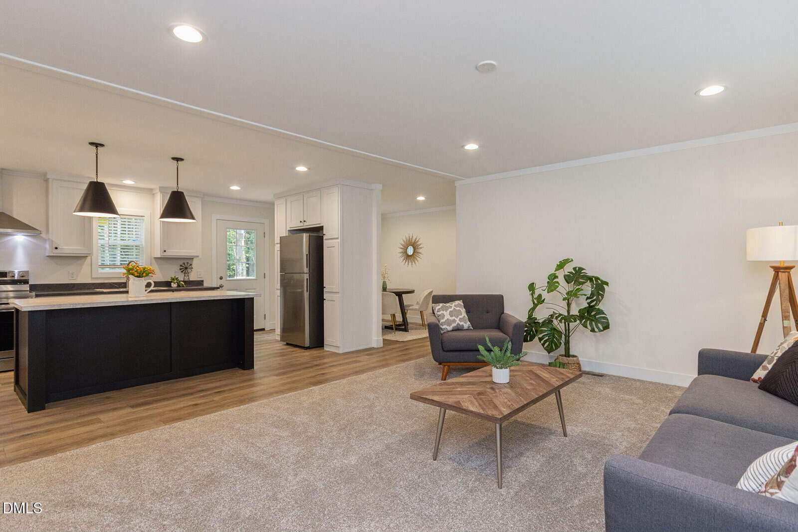 Open-concept living room and kitchen with neutral walls, recessed lighting, a gray sofa, wooden coffee table, indoor plant, modern light fixtures, stainless steel appliances, and a dining area visible in the background.