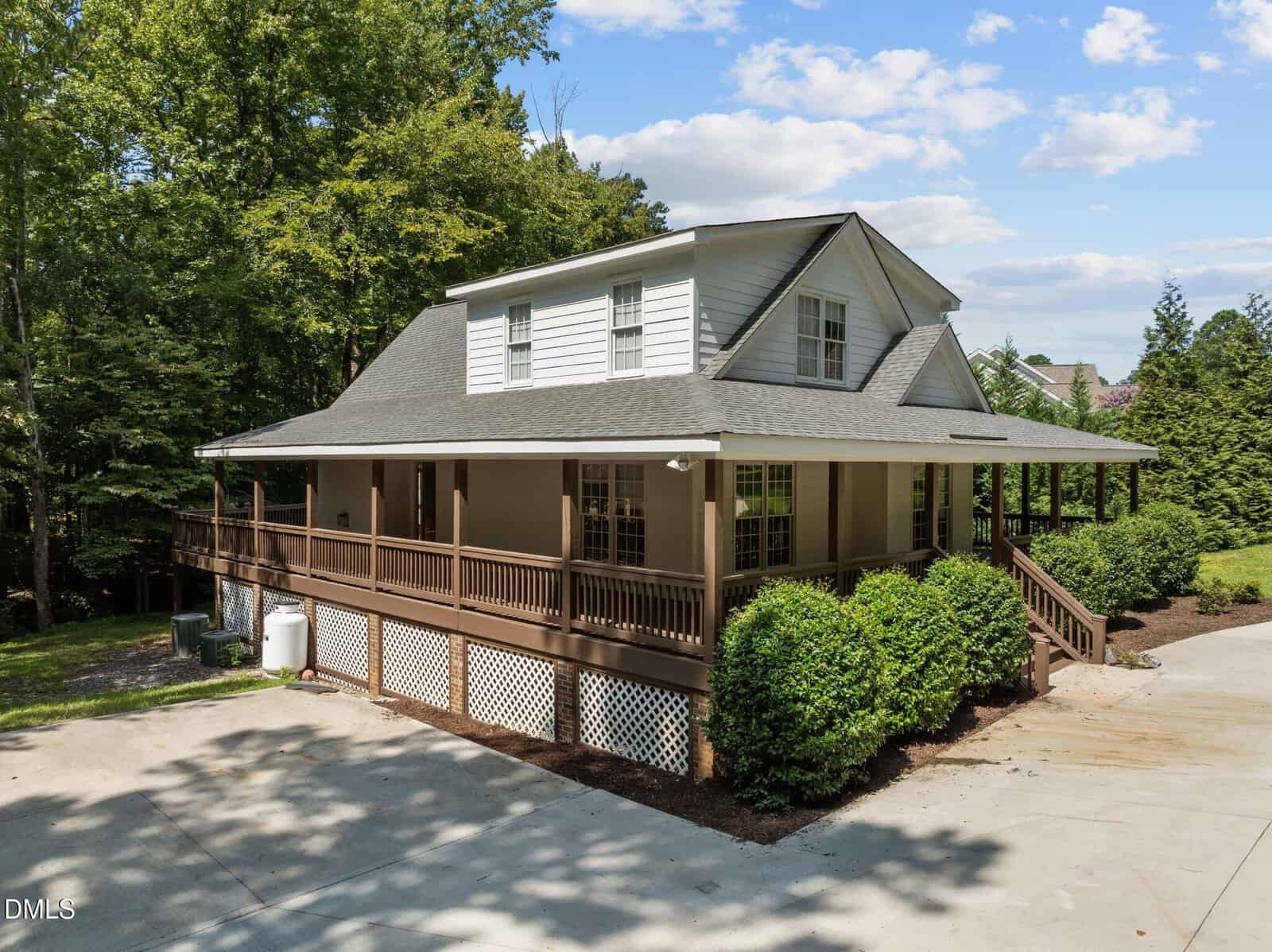 A two-story house at 287 Somerset Lane in Henderson, featuring a gray roof, white siding, large wraparound porch with brown railings, lattice under the porch, surrounded by trees and shrubs, and a concrete driveway in front.