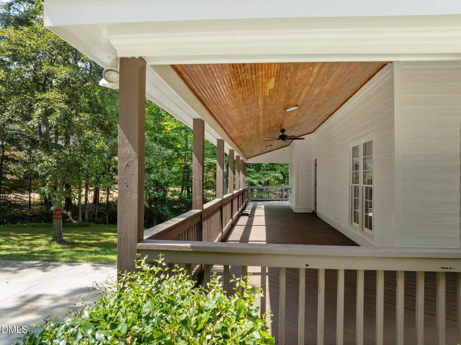 A covered wooden porch with a sloped ceiling, ceiling fan, brown railing, white siding, and large windows, overlooking a green lawn and trees in the background.