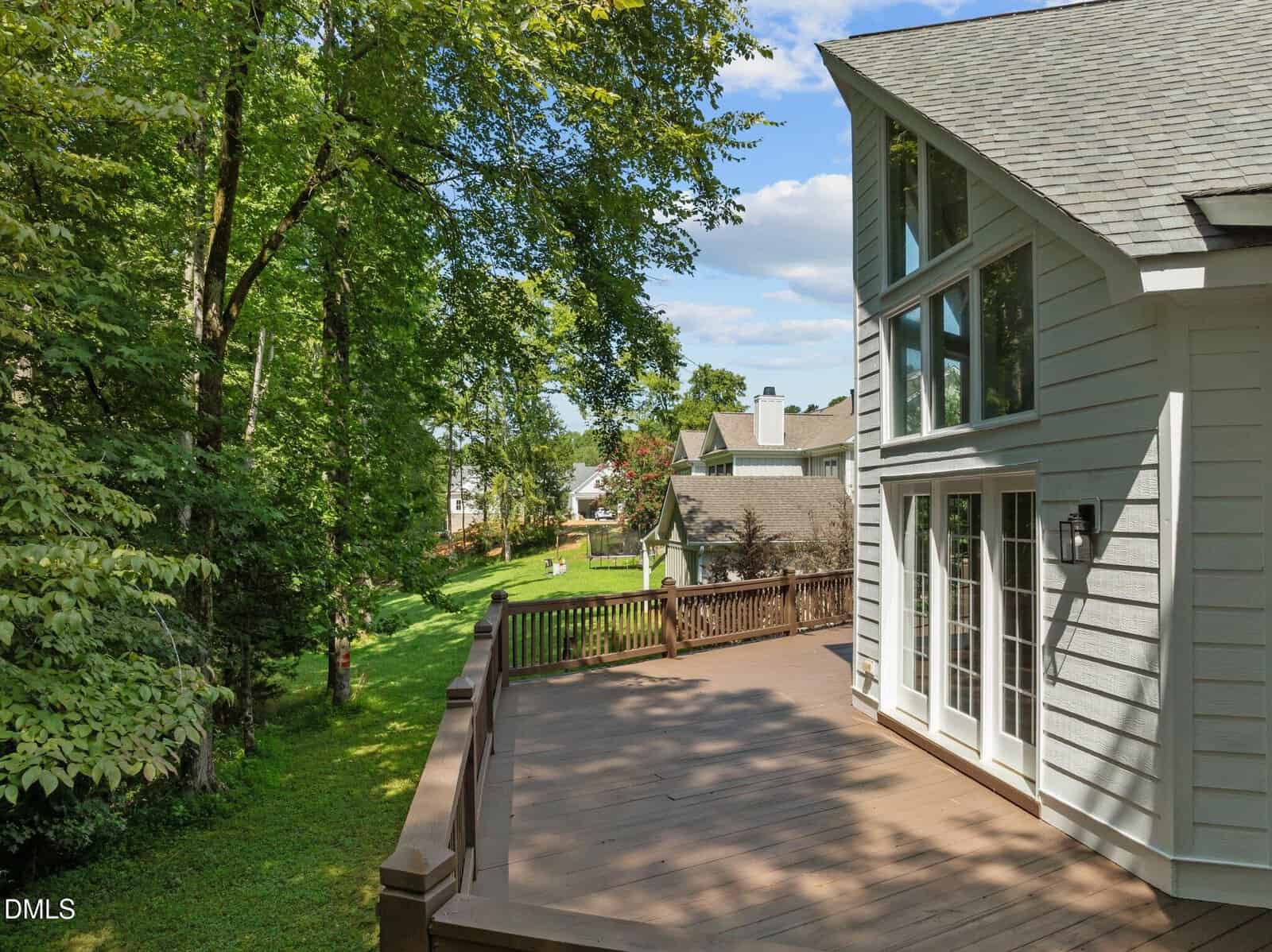 Spacious wooden deck attached to a modern white house with large windows, overlooking a grassy backyard bordered by trees and neighboring homes under a partly cloudy sky.