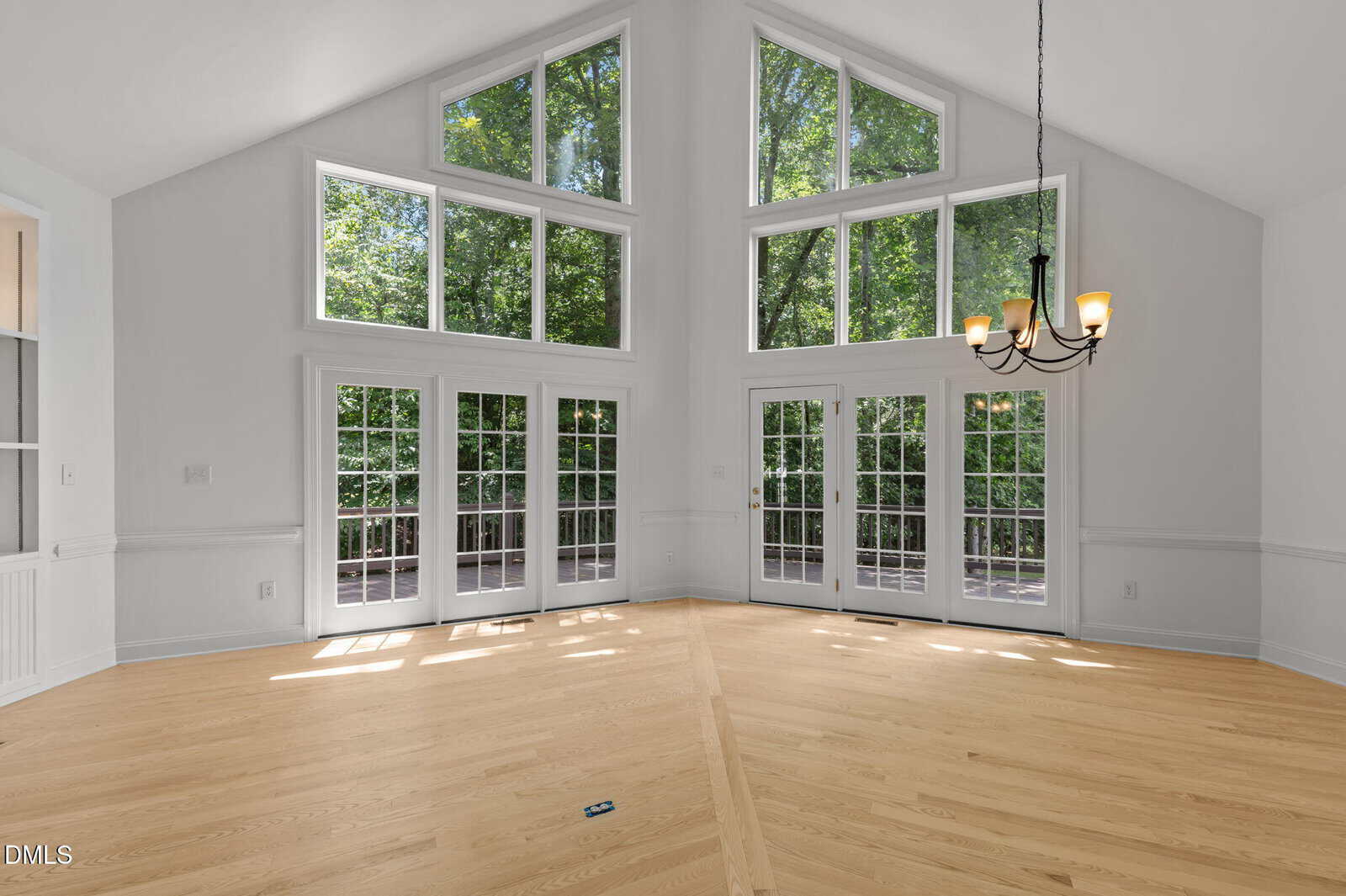 Bright, empty room with light hardwood floors, tall vaulted ceiling, large windows, multiple French doors, and a modern chandelier. Green trees are visible outside through the windows.