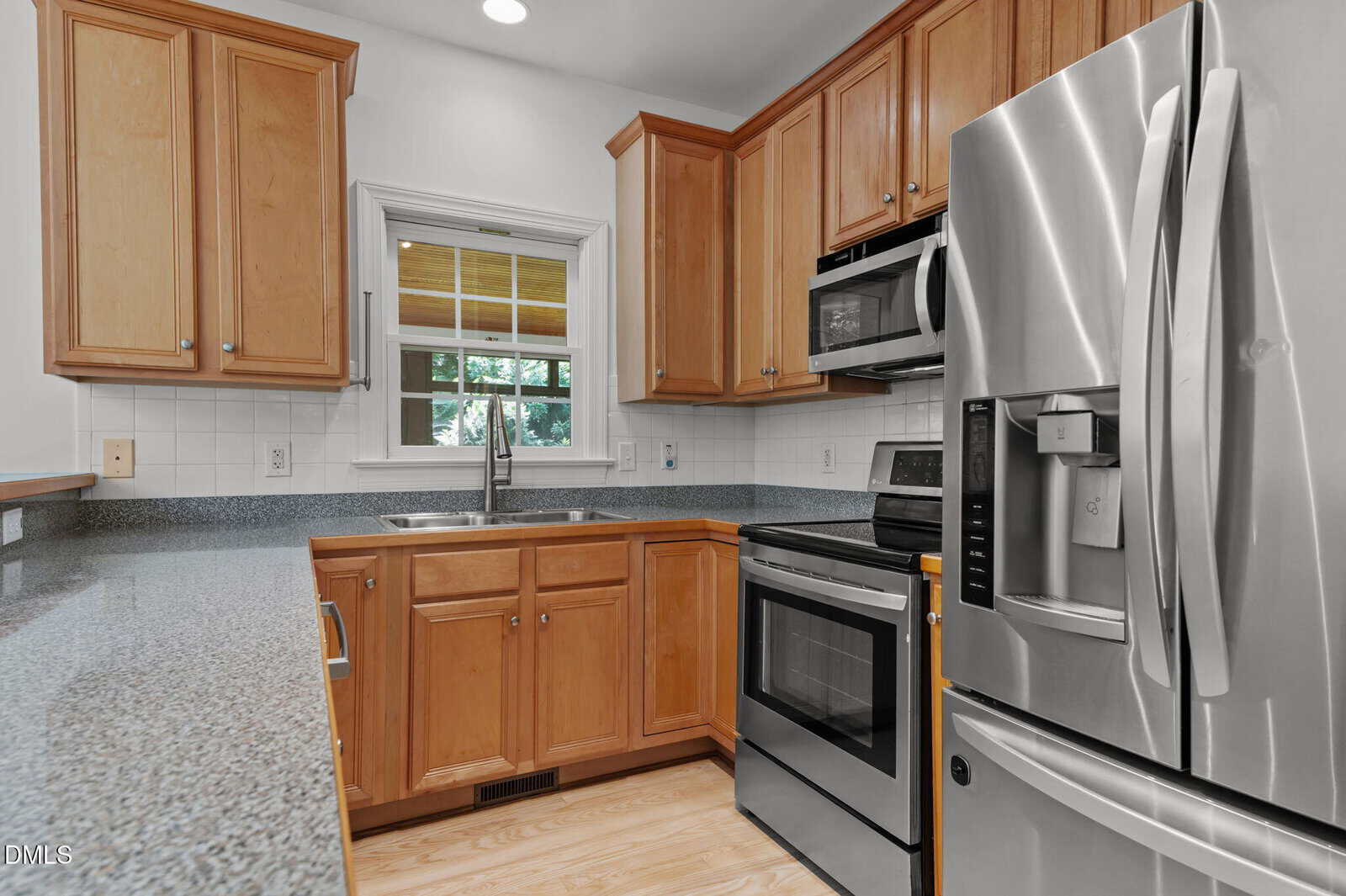 A kitchen with wooden cabinets, stainless steel appliances including a refrigerator, oven, microwave, and dishwasher, granite countertops, a double sink, and a window over the sink with white tile backsplash.