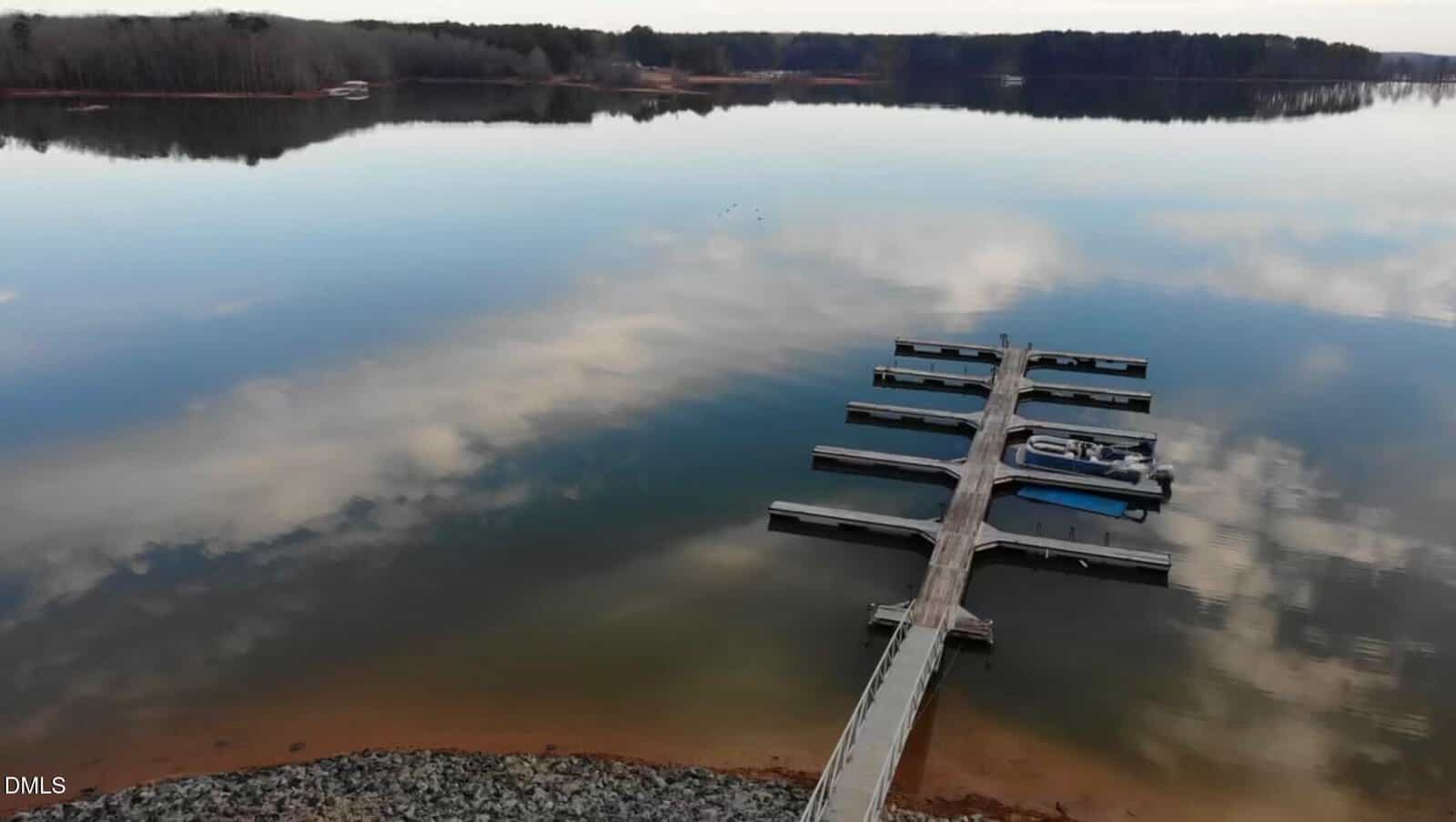 A dock extends into a calm lake with a few boats moored. The sky is reflected in the water. Trees line the far shore, and rocky ground is visible in the foreground near the dock’s entrance.