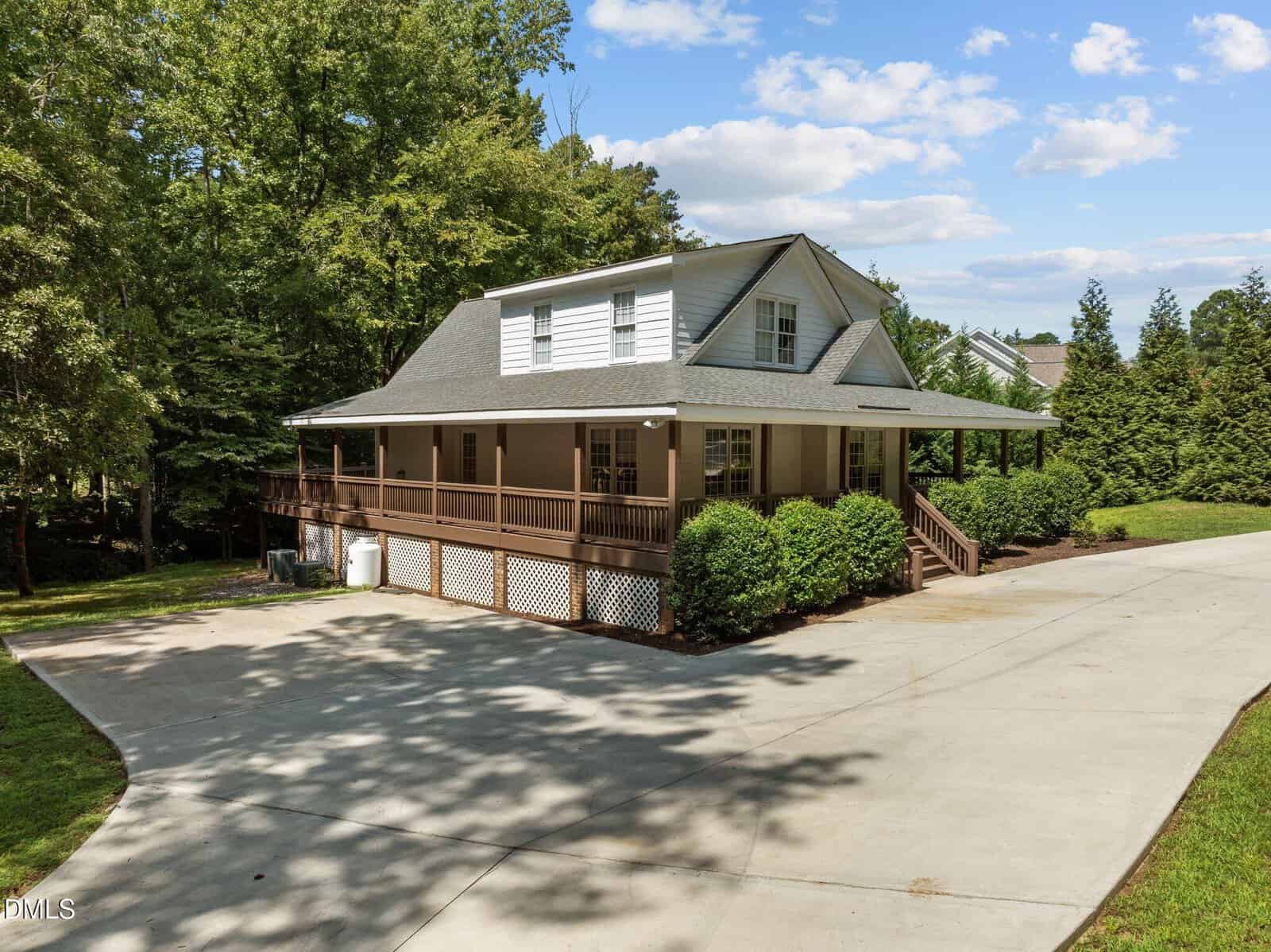 A white, two-story house with a large wraparound porch, surrounded by trees and bushes, sits next to a wide concrete driveway under a blue sky with scattered clouds.