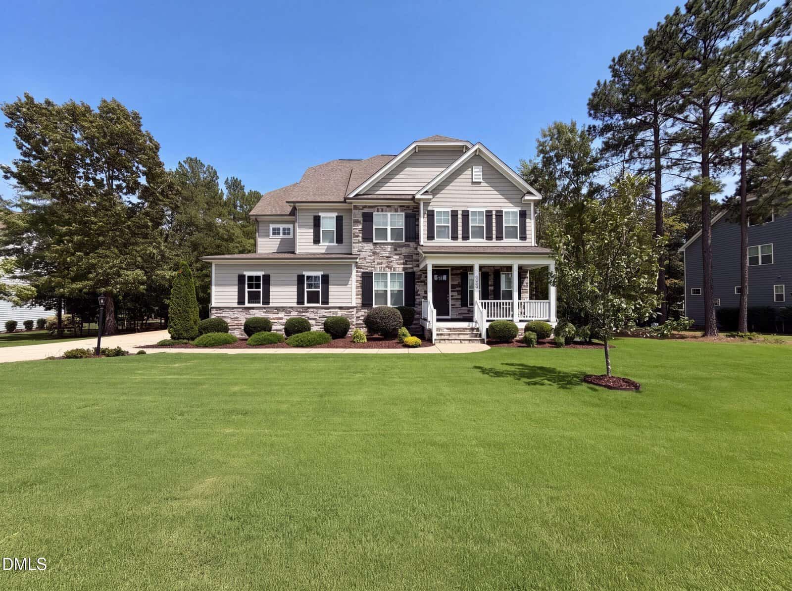 Situated at 8808 Sprouted Lane in Wake Forest, this two-story suburban house features gray siding with white trim and elegant stone accents. The home boasts a covered front porch, manicured bushes, and a large, neatly mowed front lawn dotted with trees and complemented by a driveway on the left side.
