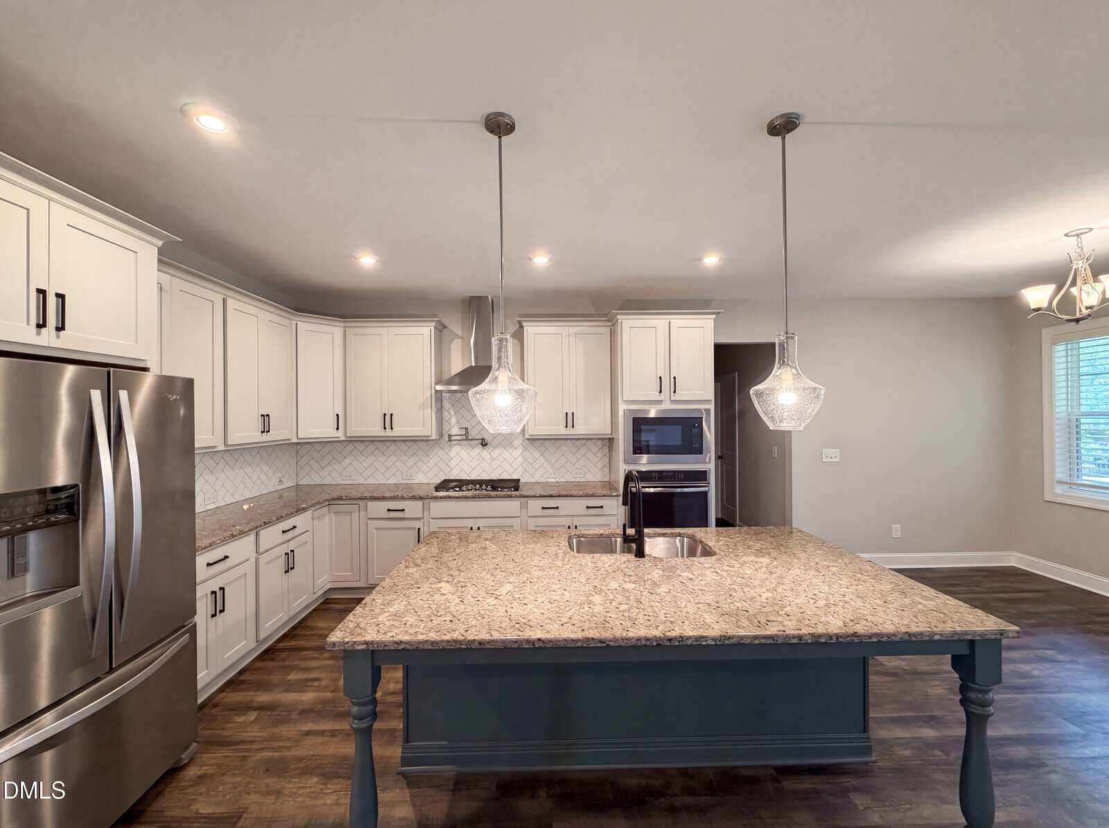 Modern kitchen with white cabinets, stainless steel appliances, granite island with sink, pendant lights, and hardwood floors. A window and a dining area are visible in the background.
