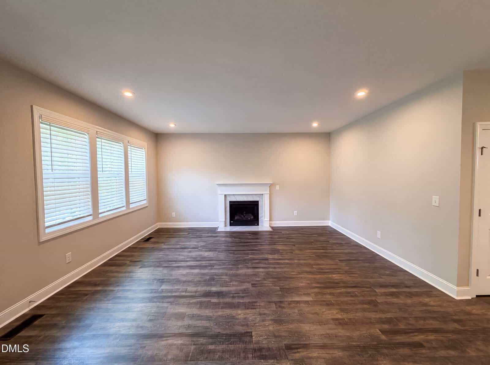 Spacious living room with wood-look flooring, three large windows on the left wall, recessed ceiling lights, and a white fireplace centered on the far wall. Walls are painted light beige. Room is unfurnished.