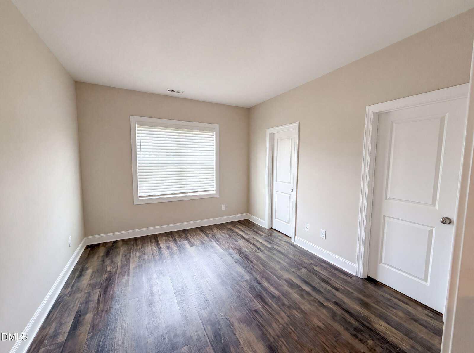 Empty room with light beige walls, a window with closed blinds, dark wood flooring, and two white doors. The room appears clean and well-lit from natural light coming through the window.