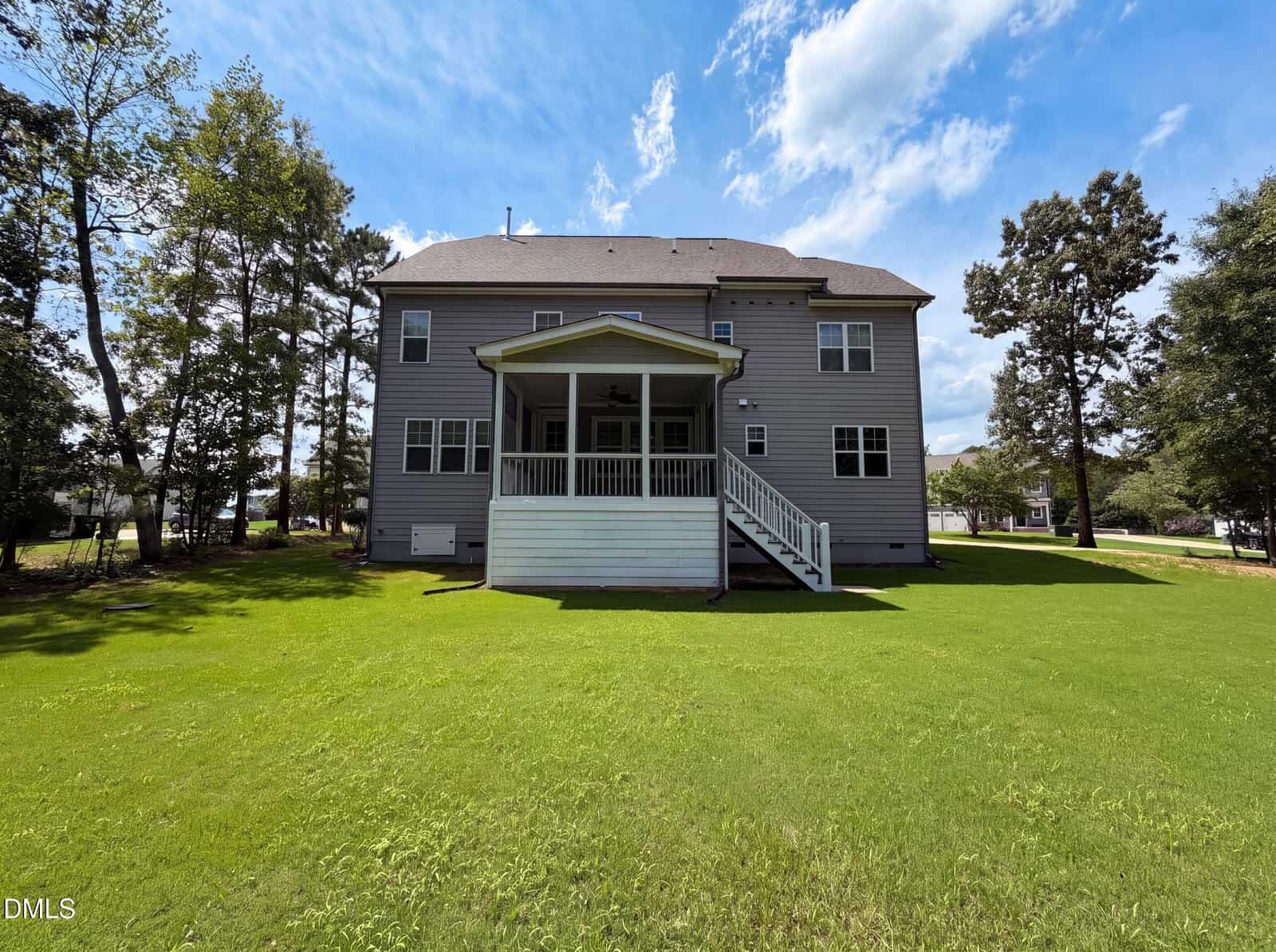 Two-story house with gray siding and white trim, shown from the backyard. A covered porch with white railings and stairs leads to a large green lawn. Several trees frame the scene under a partly cloudy sky.