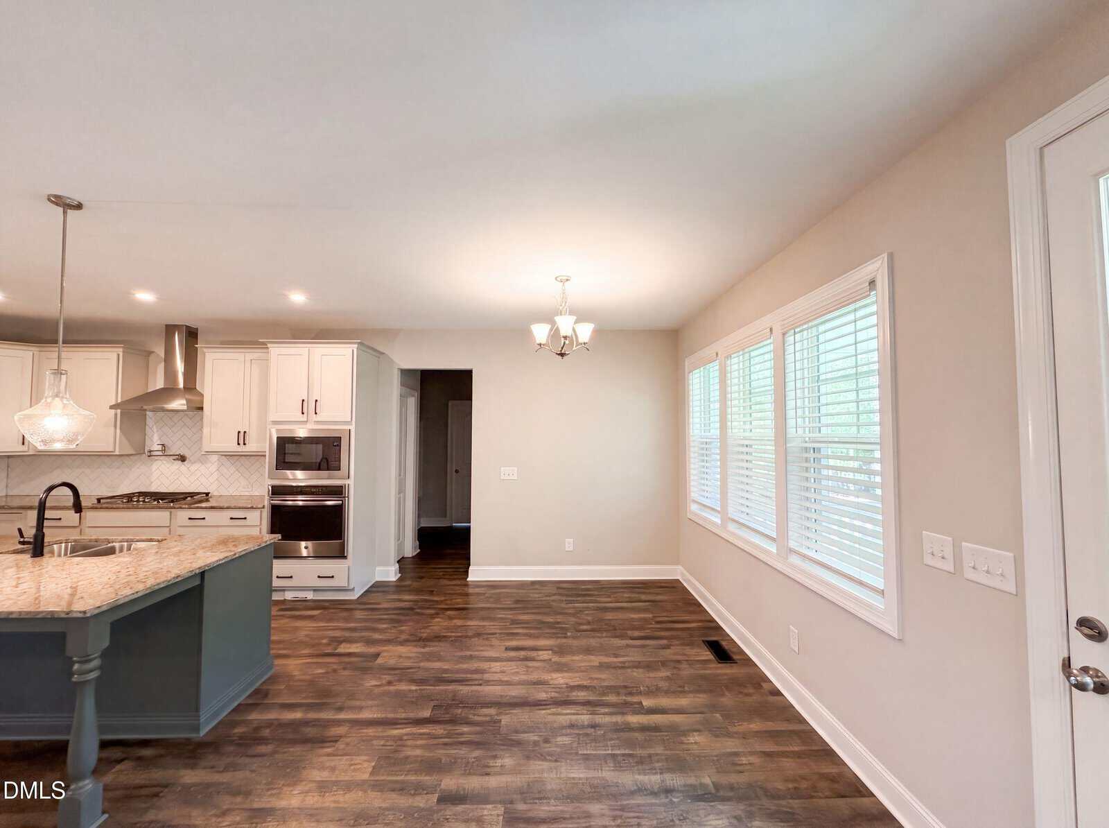 Bright kitchen and dining area with wood floors, white cabinets, stainless steel appliances, a blue island, pendant lights, chandelier, and large windows letting in natural light.