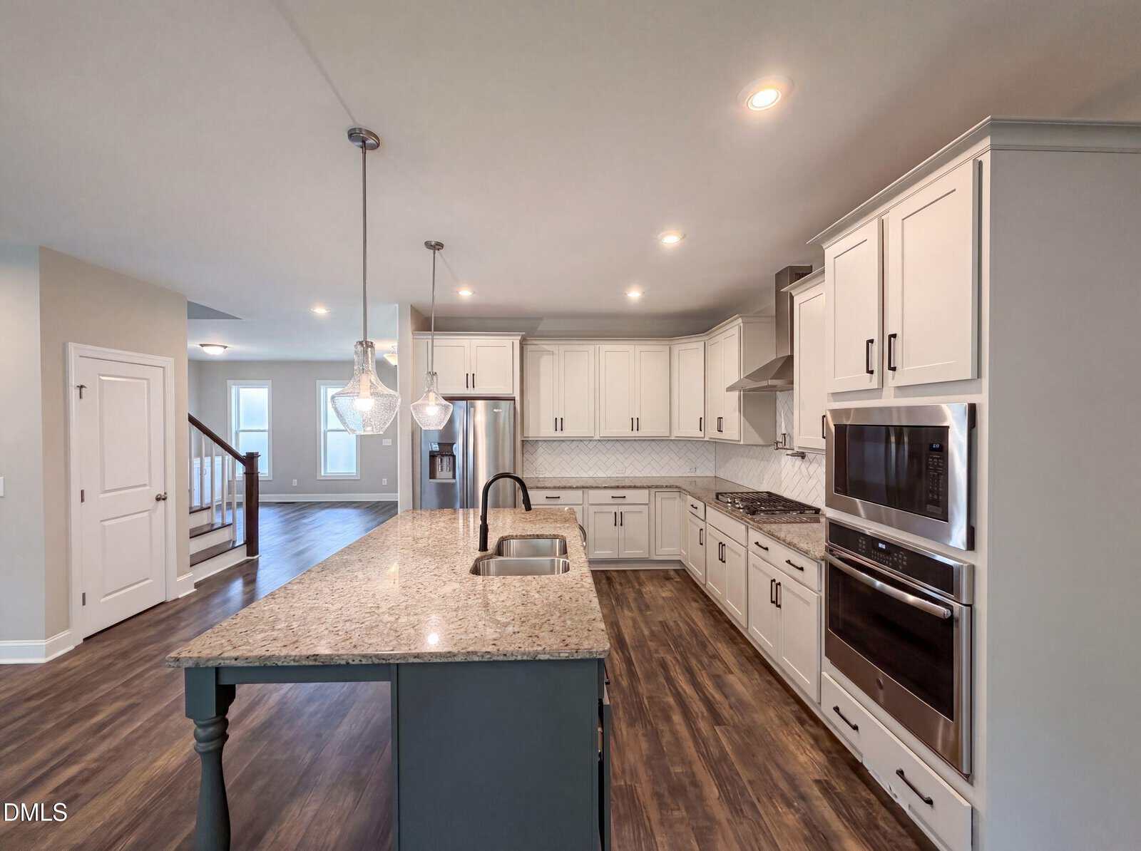 Modern kitchen with white cabinets, stainless steel appliances, granite island with sink, pendant lights, wood flooring, and a view of a staircase and adjoining room in the background.