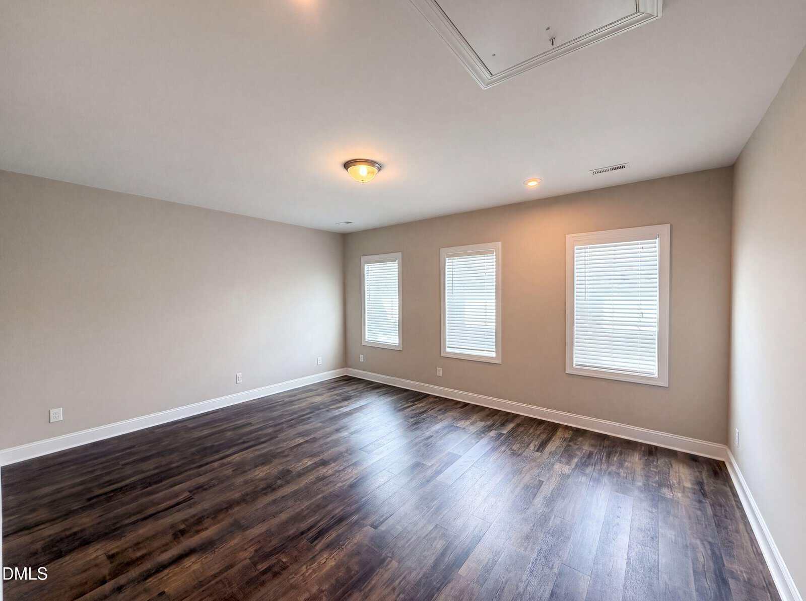 Spacious, empty room with three windows, beige walls, ceiling lights, and dark wood flooring. White baseboards line the walls.