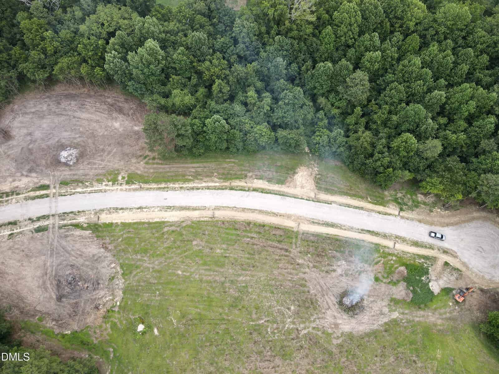 Aerial view of a curved dirt road through a partially cleared area with patches of grass, two small smoke plumes, trees bordering the scene, and a white car parked on the right side of the road.