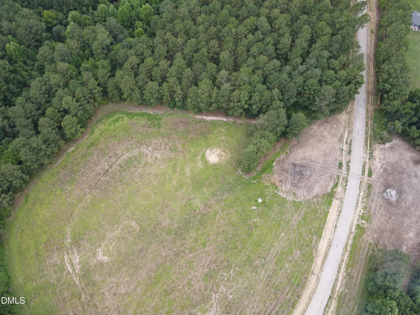 Aerial view of a partially cleared plot of land bordered by dense trees, with a dirt road curving beside it and patches of grass and bare soil visible throughout the area.