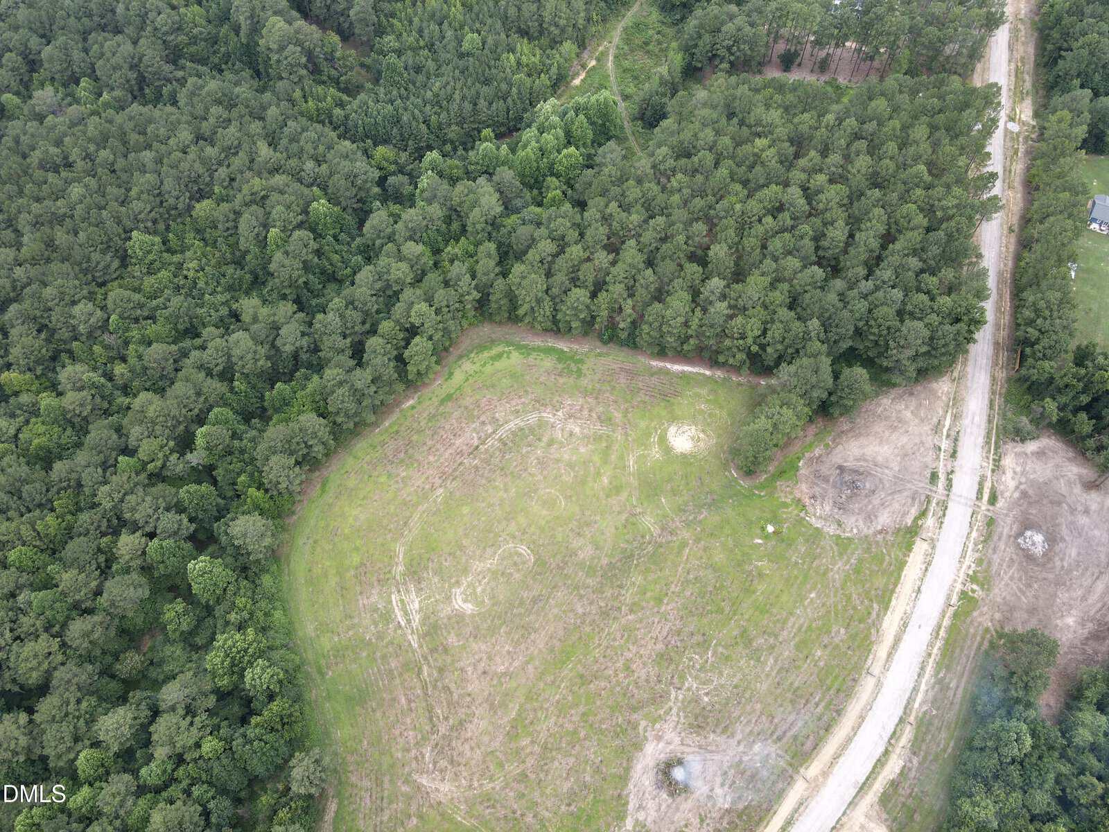 Aerial view of a cleared grassy area surrounded by dense trees, with a dirt road running along the right side. Some patches of bare soil and forest border the open space. A few scattered structures are visible near the road.