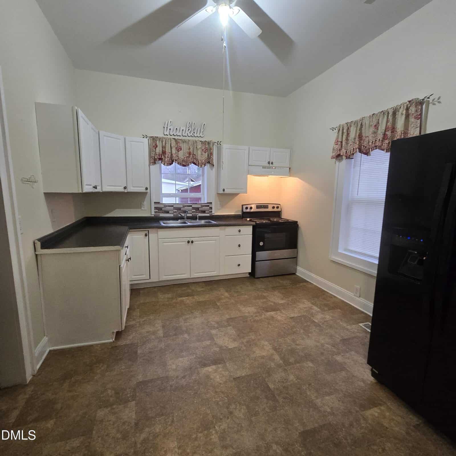 Kitchen with white cabinets, black countertops, stainless steel stove, black refrigerator, window with floral valance, ceiling fan with light, and "thankful" sign above sink. Brown patterned floor and neutral walls.
