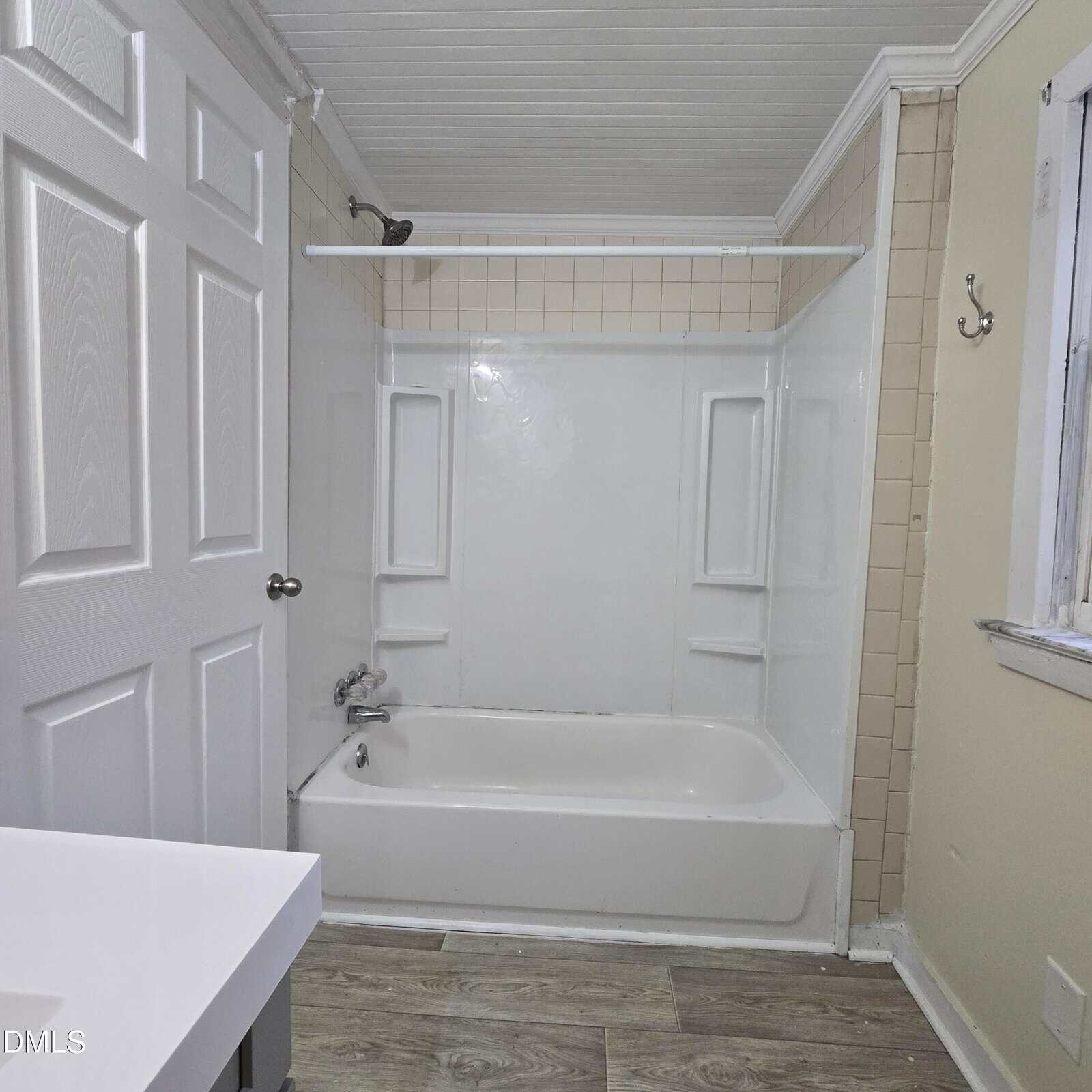 A bathroom with a white bathtub and shower, beige tile walls, a white door, a wall-mounted shelf, a window, and wood-look flooring. The tub has a curtain rod and built-in shelves on both sides.