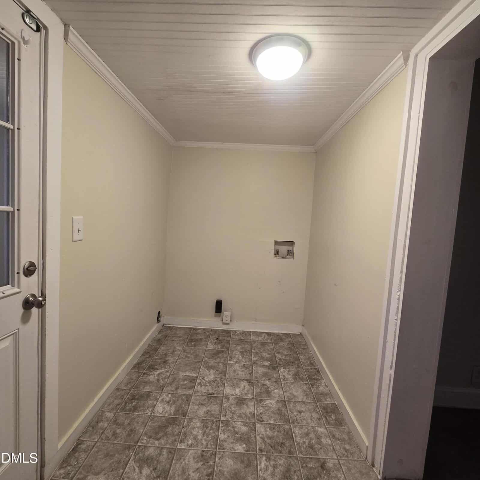 Small empty laundry room with beige walls, gray patterned tile flooring, a ceiling light, wall outlets, and washing machine hookups on the back wall; door with glass window on the left.