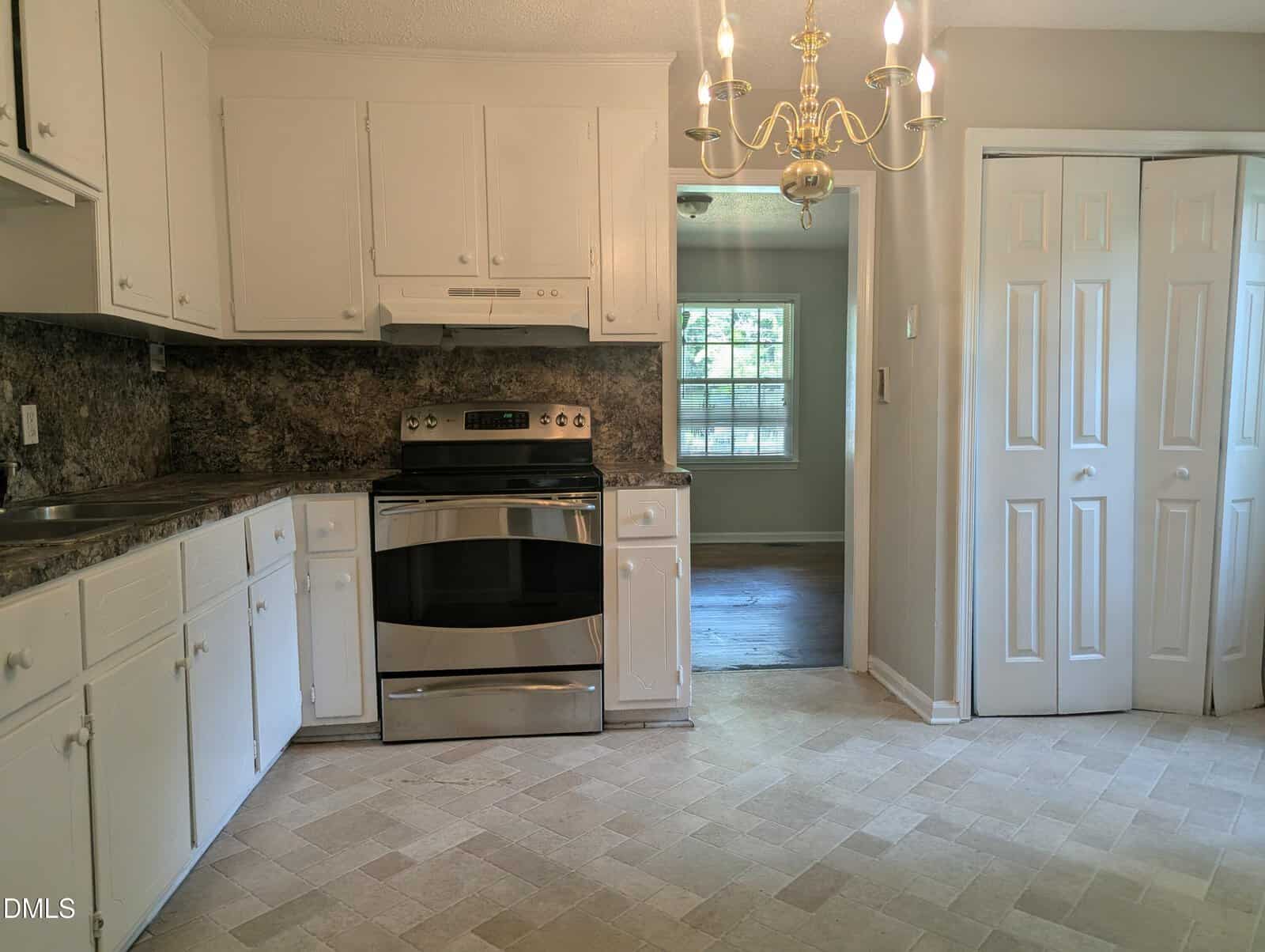 A kitchen with white cabinets, stainless steel oven, marble-patterned countertops, chandelier light fixture, and folding closet doors. An adjacent room with wooden flooring and a window is visible in the background.
