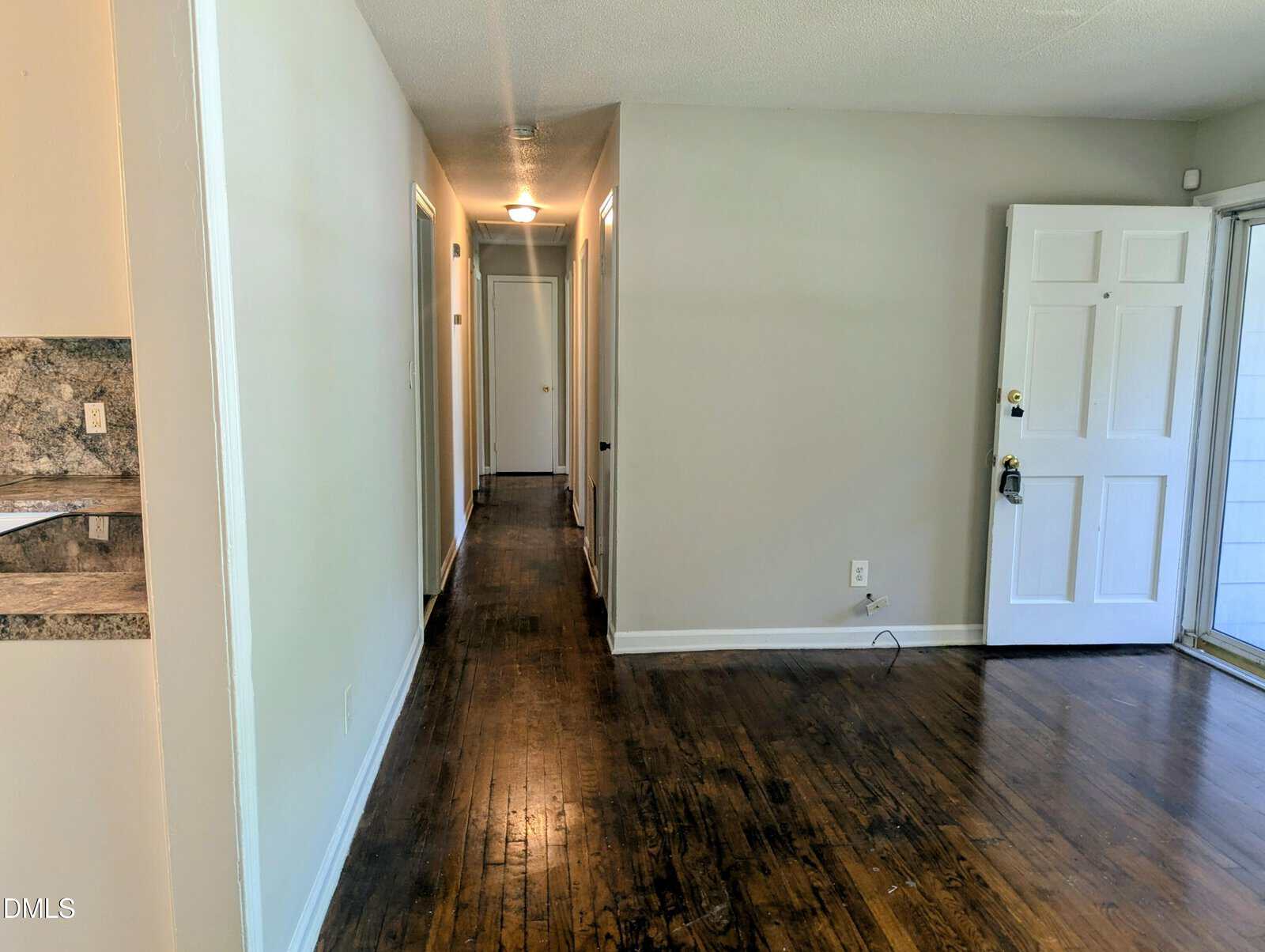 A hallway with white walls and dark wood floors extends toward the back, with several doors on each side. An open white front door is on the right, and part of a marble-patterned wall is visible on the left.