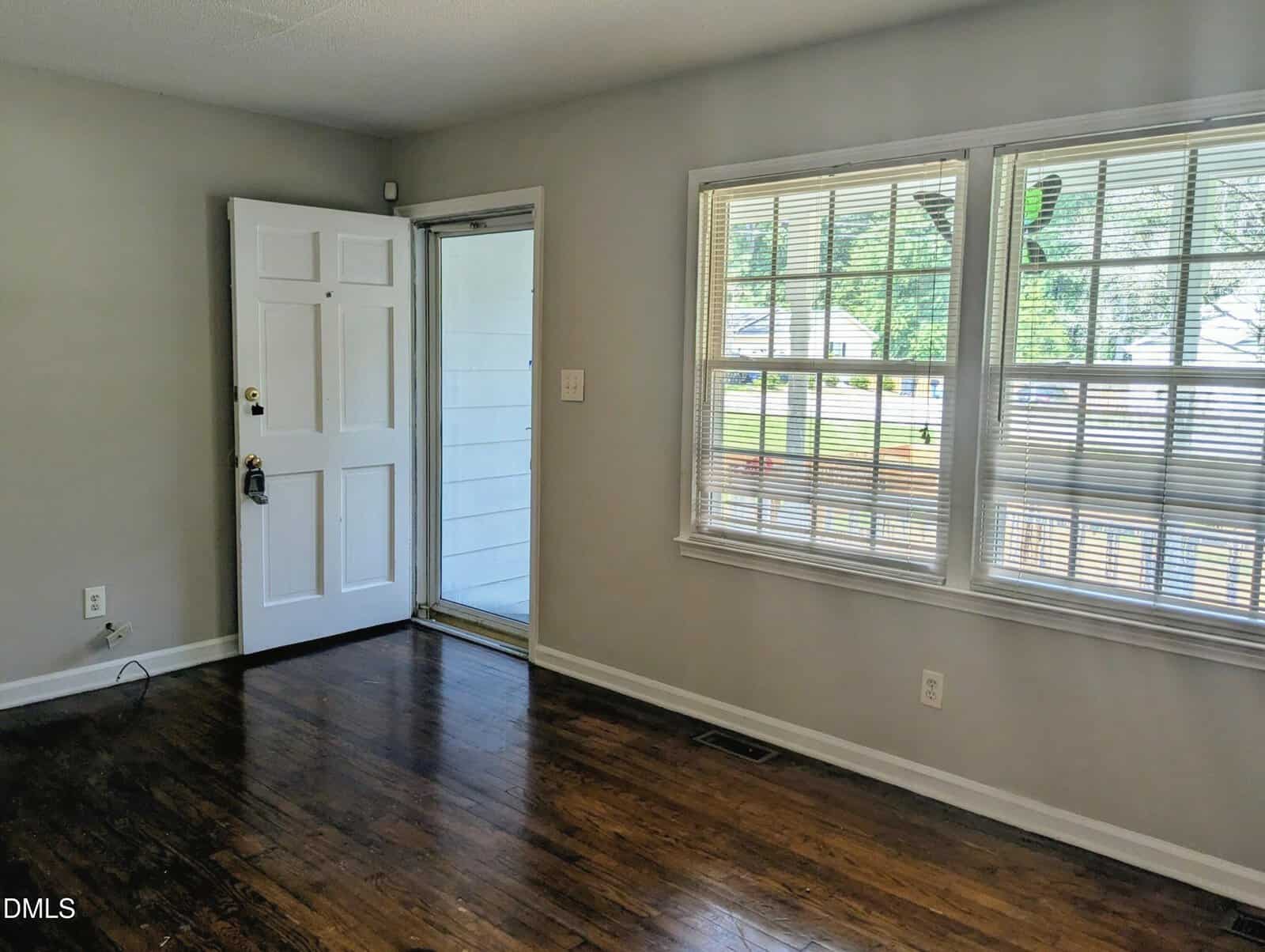 A small, empty room with light gray walls, a white front door, and two large windows with blinds. Dark wood floor and some visible electrical outlets. Sunlight is streaming in from outside.