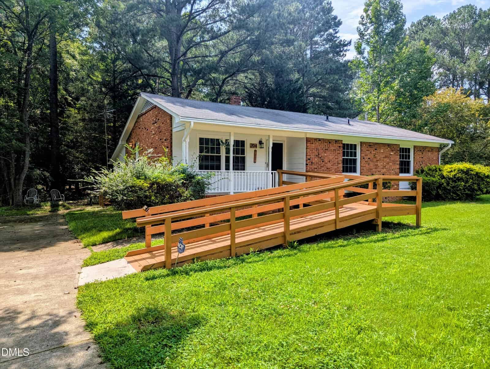 Single-story brick house with a white porch and a wooden wheelchair ramp leading to the front door. The yard is grassy with bushes and trees in the background. A concrete path runs beside the ramp.