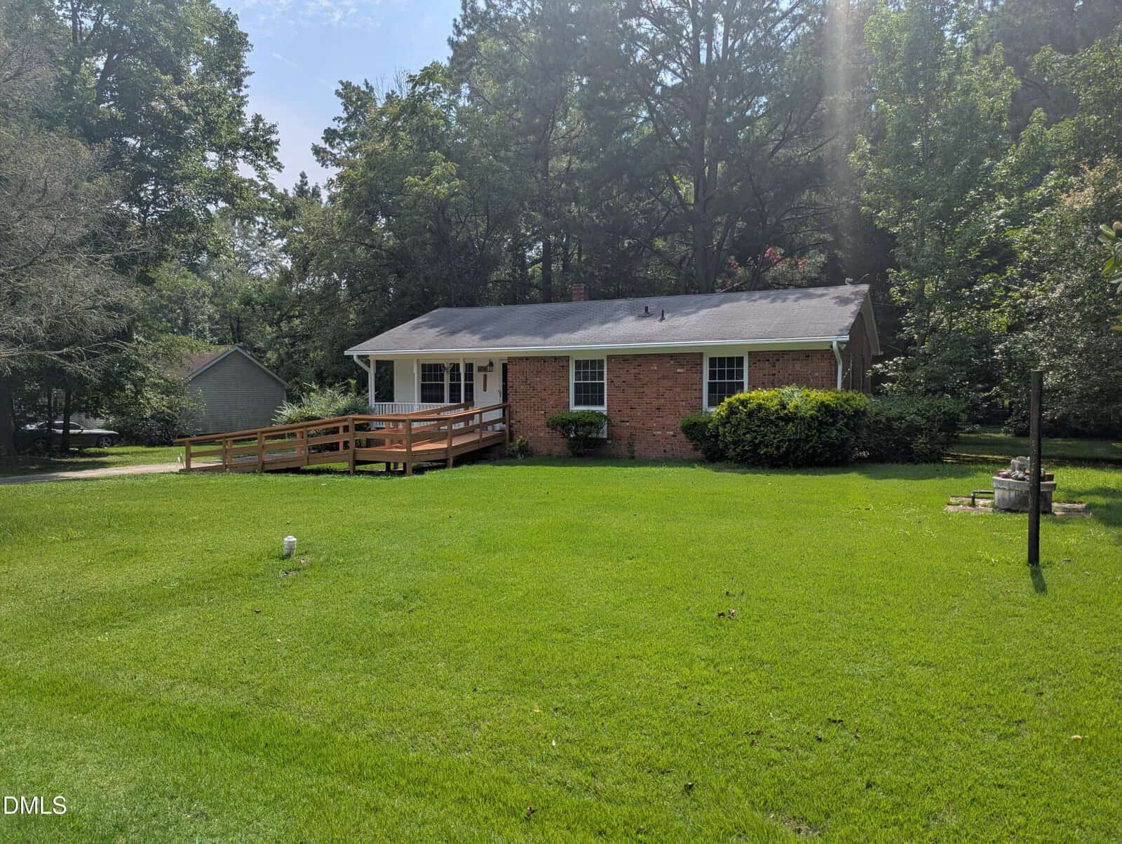 A one-story red brick house with a ramp leading to the front porch sits on a well-maintained lawn surrounded by trees, with a driveway and a small shed visible in the background.