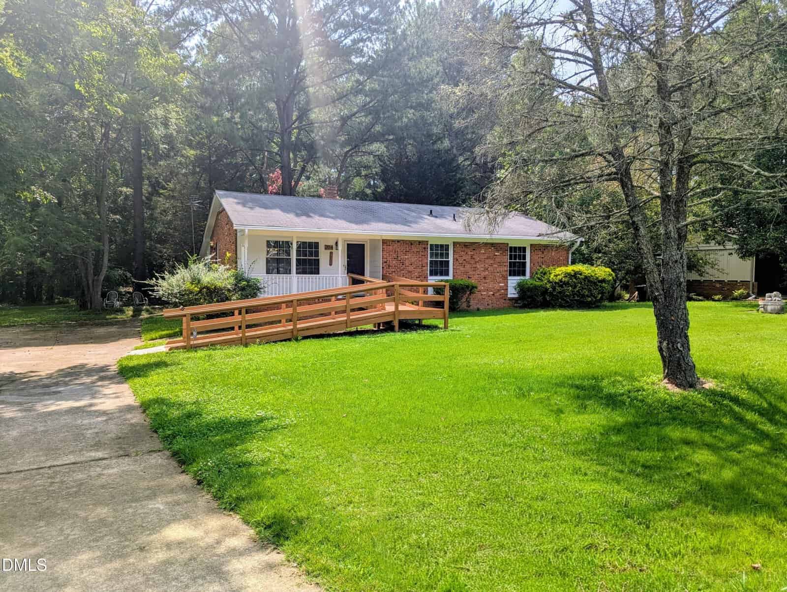 A single-story brick house with white trim at 204 Drew Street in Louisburg features a wooden wheelchair ramp leading to the front porch. The home is surrounded by a green lawn and trees, with a concrete driveway visible in the foreground.
