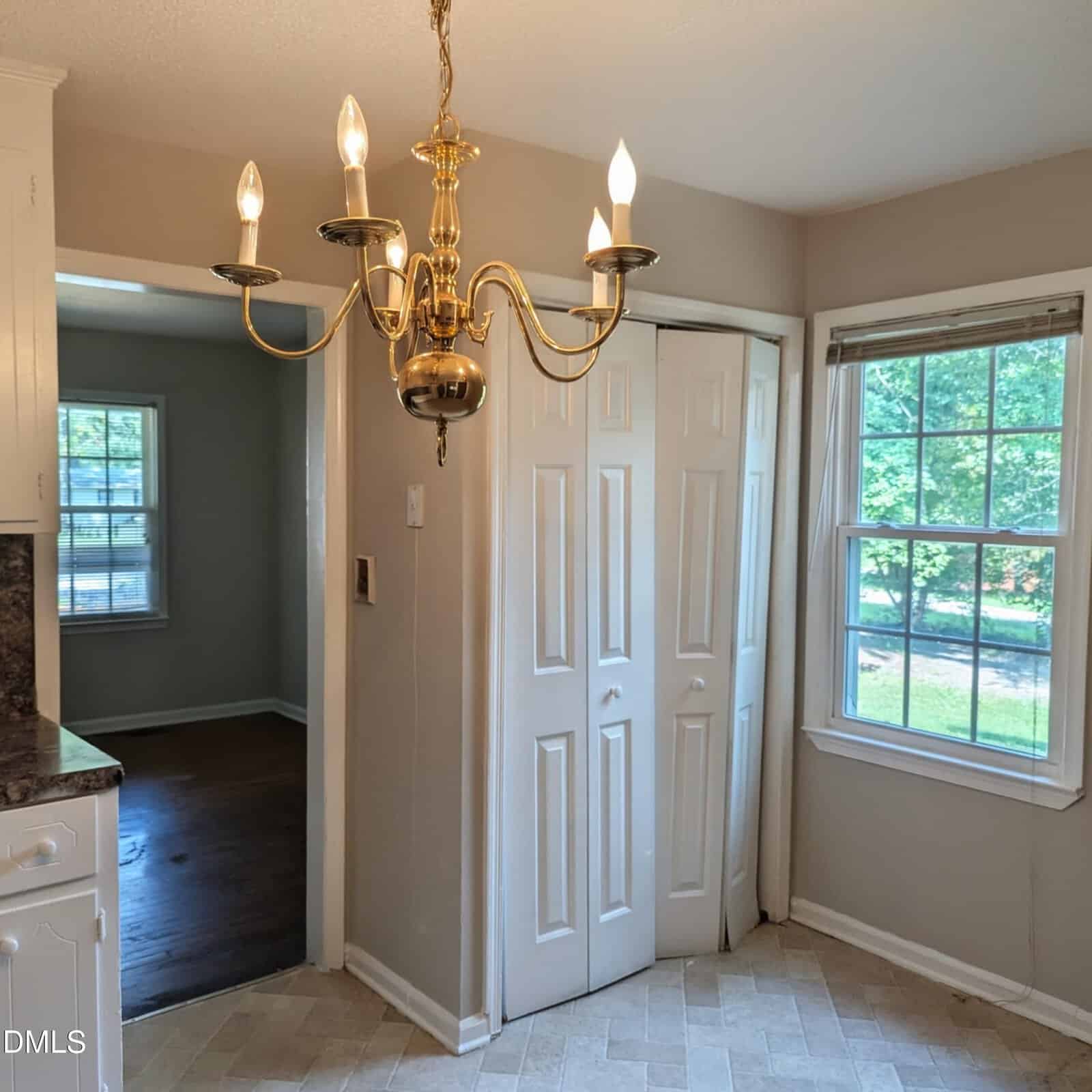 A kitchen corner with a gold chandelier, beige walls, white folding pantry doors, a window with a view of greenery, and an adjoining room with dark wood flooring.