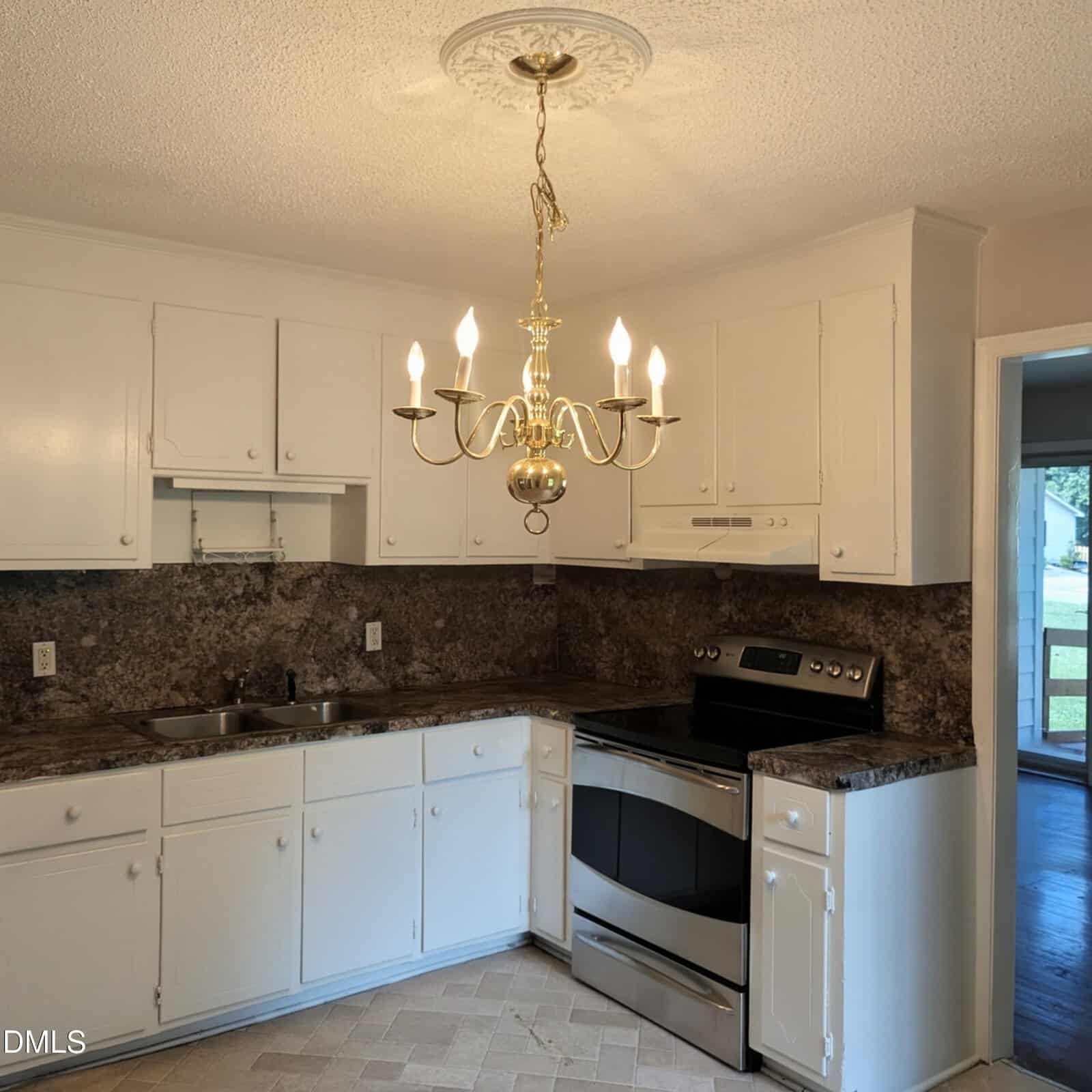 A kitchen with white cabinets, dark granite countertops, a stainless steel stove, double sink, and a gold chandelier. A doorway opens to a room with large windows and a view outside.