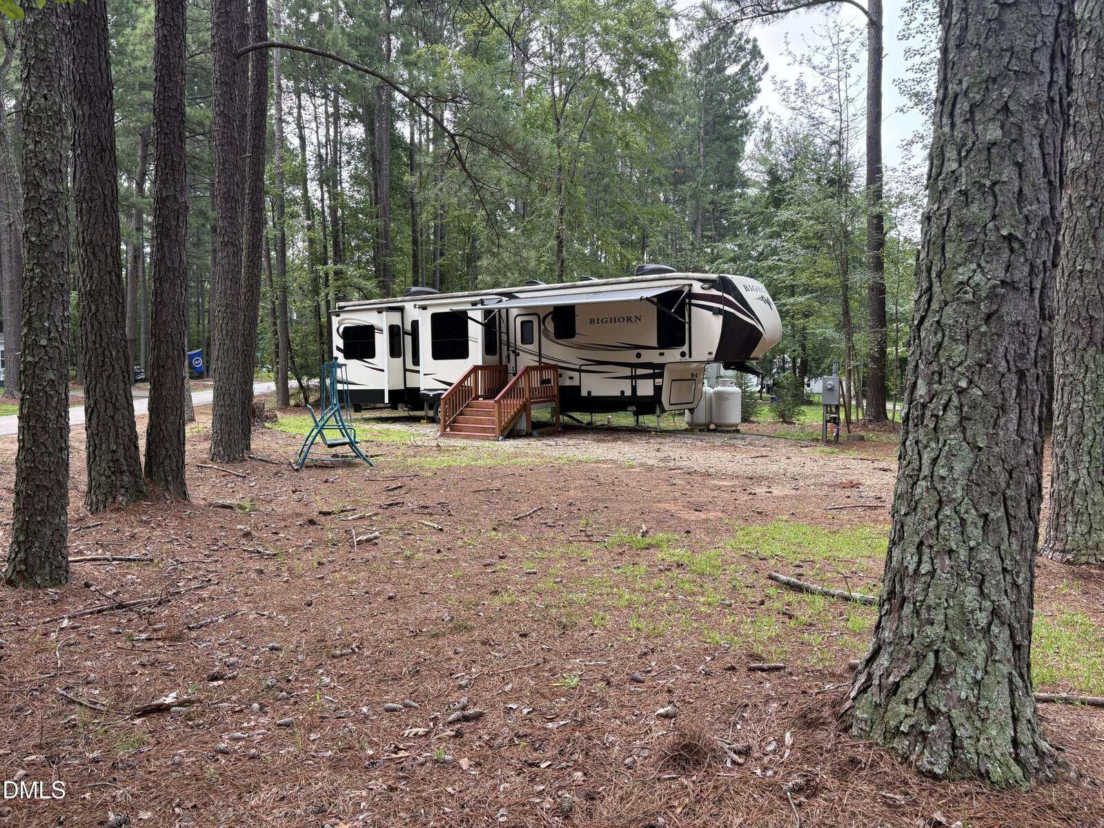 A large fifth-wheel camper is parked among tall pine trees on a dirt lot. Wooden steps lead to the door, with propane tanks visible in front. A blue-and-green swing set sits to the left of the trailer.